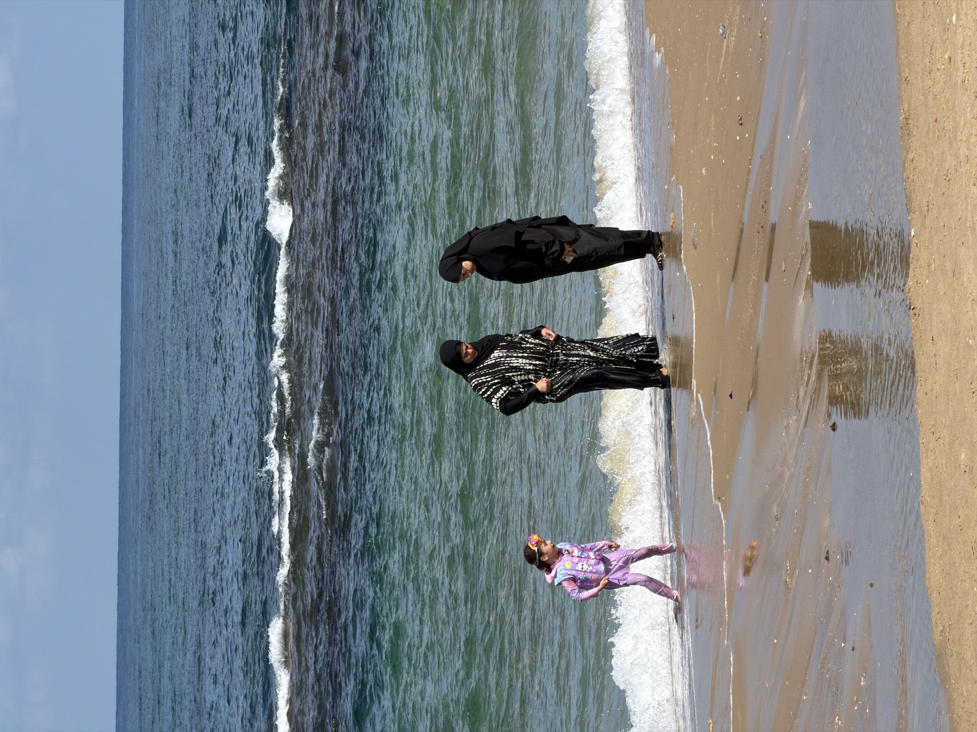In Tel-Aviv, Israel, two women and a young girl are by the sea. The image is taken from a high angle, looking down. The two women are dressed in Islamic attire, with one wearing a black abaya and hijab, and the other a black and white patterned abaya with a hijab. The young girl is wearing a purple swimsuit with a cartoon design. She has goggles on her head. They are standing on a sandy beach with waves from the sea washing up to the shore. The water is a deep blue-green, and the sky is a light blue. The sun is shining, creating reflections on the wet sand. The scene suggests a pleasant day at the beach, possibly in the afternoon given the light.