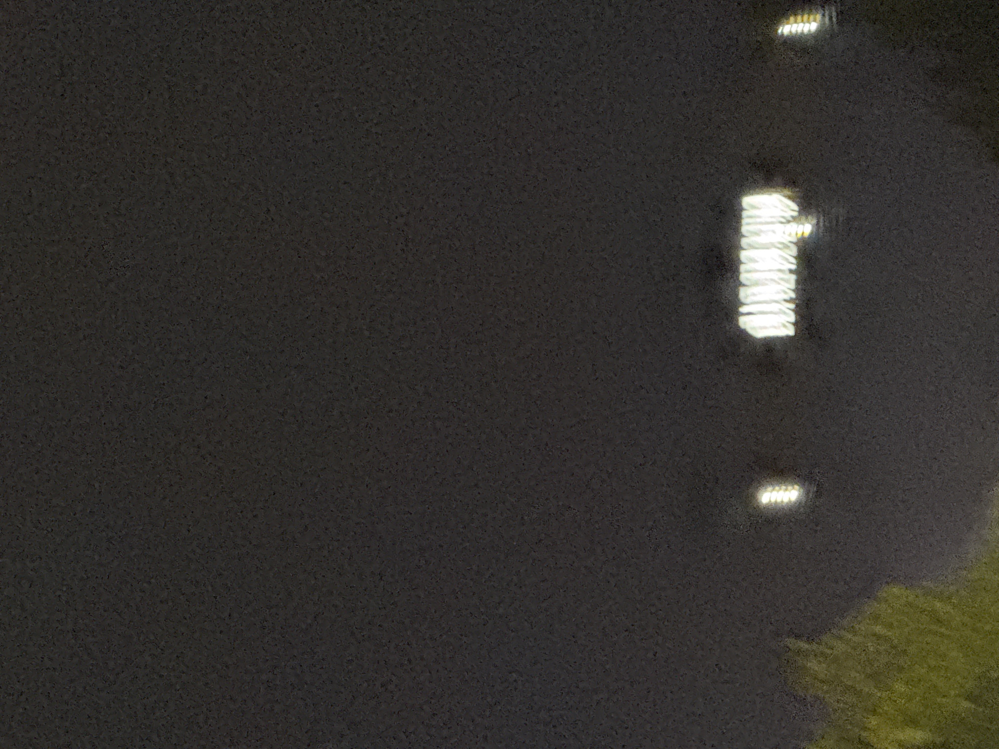 The image captures a nighttime view of Tbilisi, Georgia, with a dark, grainy sky dominating the frame. In the lower portion of the image, a blurred section of green foliage is visible on the left, likely trees. Further to the right, two distinct clusters of lights are present. The left cluster consists of a single bright light. The central cluster is a larger, elongated rectangular shape composed of multiple bright lights arranged horizontally, possibly indicating a building or a sign. To the far right, another vertical arrangement of bright lights is faintly visible. The overall impression is one of urban lights against a dark night sky, with a hint of natural elements in the foreground.