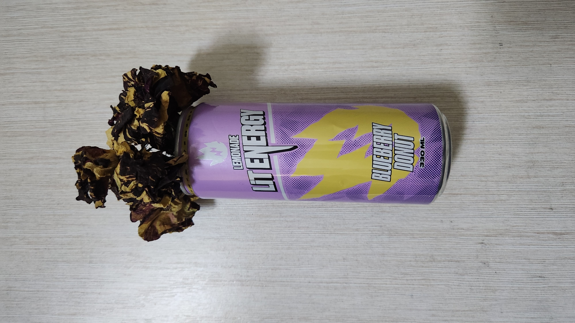 A still life photograph shows a can of "LIT ENERGY" beverage with a "BLUEBERRY DONUT" flavor, with dried flowers on top, resting on a light-colored wooden surface. The can is predominantly purple with yellow lightning bolt graphics. The background is a simple, light-colored wooden wall or table with a visible wood grain texture. There is no indication of the city of Buynaksk, Russia, in the image.