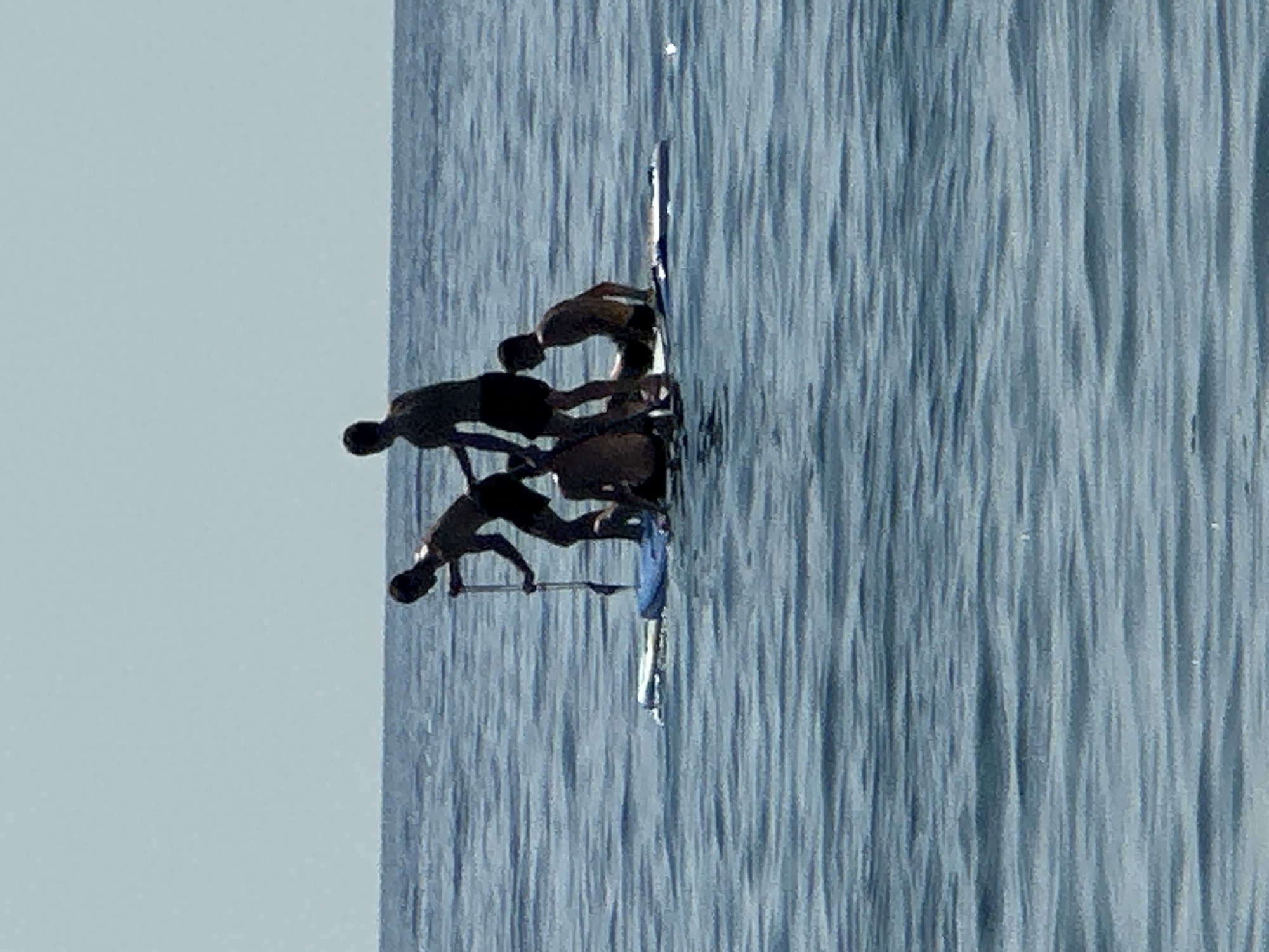 Four young males are on a paddleboard in the calm, blue sea under a clear sky. The paddleboard is partially submerged in the water, with a blue inflatable pad on top. One male is standing and holding a paddle, appearing to be paddling. Another male is standing behind him, and two other males are sitting on the paddleboard. The scene is outdoors, and the lighting suggests it is daytime. The location is identified as Đurovići, Montenegro.