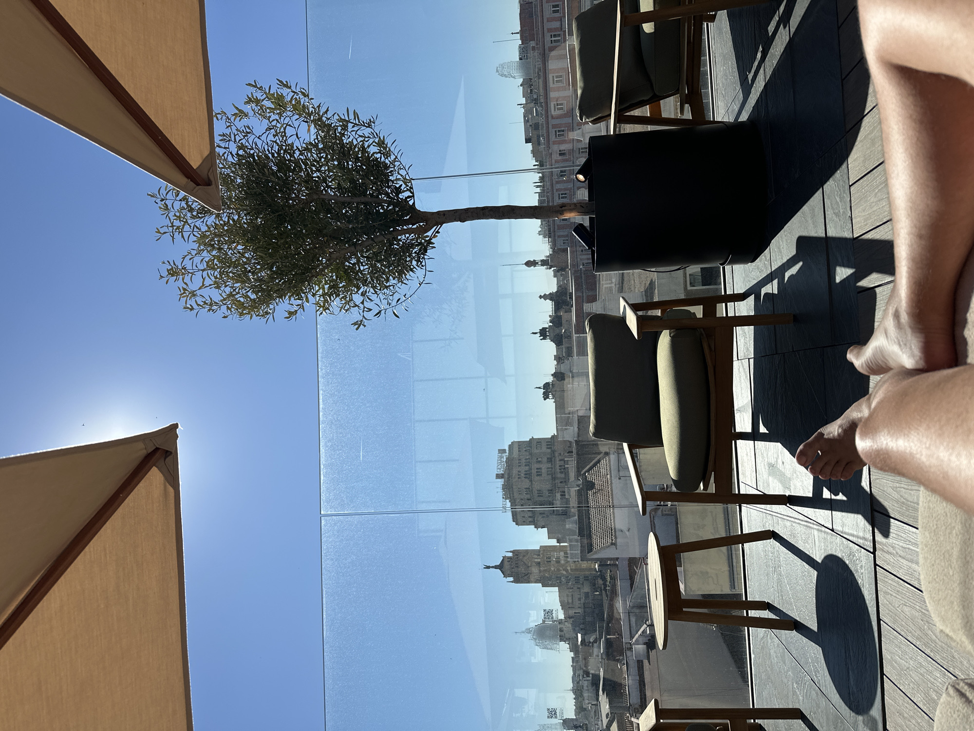The image, taken at a high angle, shows an outdoor seating area, possibly on a rooftop terrace, with a view of Madrid, Spain. The day is sunny, casting harsh shadows on the decking floor. Beige-colored parasols are partially visible on the top and bottom left. In the center, a potted olive tree stands behind a glass barrier, partially obscuring the cityscape view. Reflections in the glass show buildings, towers, and domes that are characteristic of Madrid. Modern outdoor furniture, including chairs and a small table, is arranged on the terrace. At the right edge, parts of a person's legs and feet are visible, resting on a seating cushion, suggesting someone is relaxing in the area.