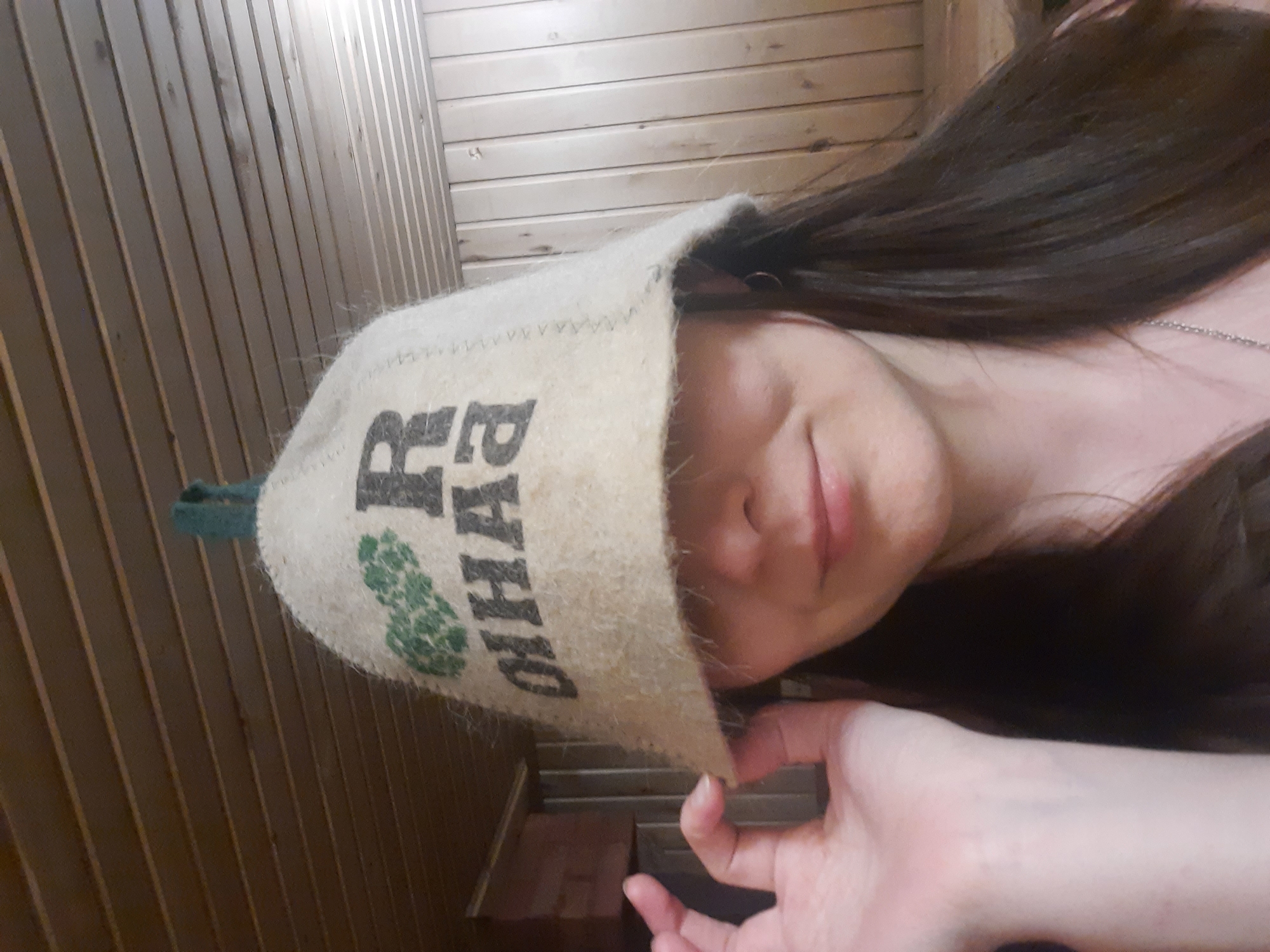 A person is wearing a sauna hat with text in Russian and a green design. They are likely in a sauna or a room with wood paneling. The image was taken from a slightly upward angle. There is no information that verifies that the person is in the city of Krasnogorskoye, Russia.