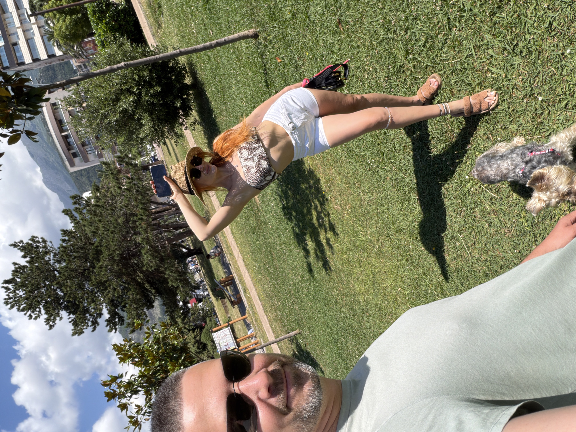 A wide-angle outdoor selfie captures two individuals and a dog relaxing on a sunny day in what appears to be a park or grassy area. The image is oriented sideways, with the man taking the selfie visible in the bottom right portion of the frame (when viewed upright).
In the foreground, an adult man is partially visible, featuring his head and shoulders. He has short dark hair, a neatly trimmed beard, and wears dark sunglasses and a light green t-shirt. He appears to be smiling subtly.
In the mid-ground, an adult woman is lying on her back on the grass. She has long red hair, is wearing a straw hat and dark sunglasses, a patterned bandeau-style crop top, and white shorts. Her legs are adorned with multiple anklets. She holds a dark-colored smartphone in her left hand. Near her hips, a black pouch with pink accents is visible, featuring the text "COVERI".
Next to the woman's feet, a small, shaggy dog, possibly a terrier mix, is also lying on the grass, wearing a red harness.
The background reveals a picturesque setting. Lush green trees and foliage are scattered across the scene. Beyond the trees, several multi-story buildings are visible, suggesting an urban or resort area. Further in the distance, large mountains rise under a sky dotted with white clouds. A wooden pole lies on the grass in the mid-ground, and a wooden bench or picnic table can be seen further back.
The activity appears to be casual relaxation outdoors. The bright sunshine, clear sky, and relaxed postures of the subjects indicate pleasant weather. The setting, with mountains and buildings, suggests a scenic location.