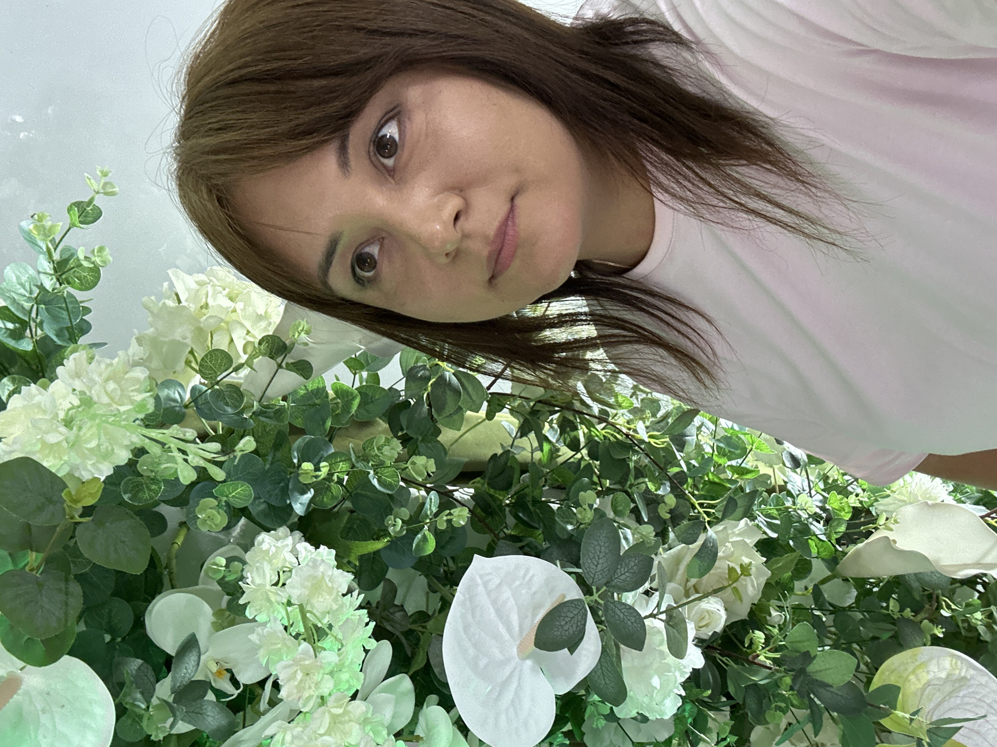A woman with light brown hair and brown eyes is photographed from a low angle, with a background of white flowers and green leaves. She is wearing a white t-shirt. The location is Unknown, Unknown.