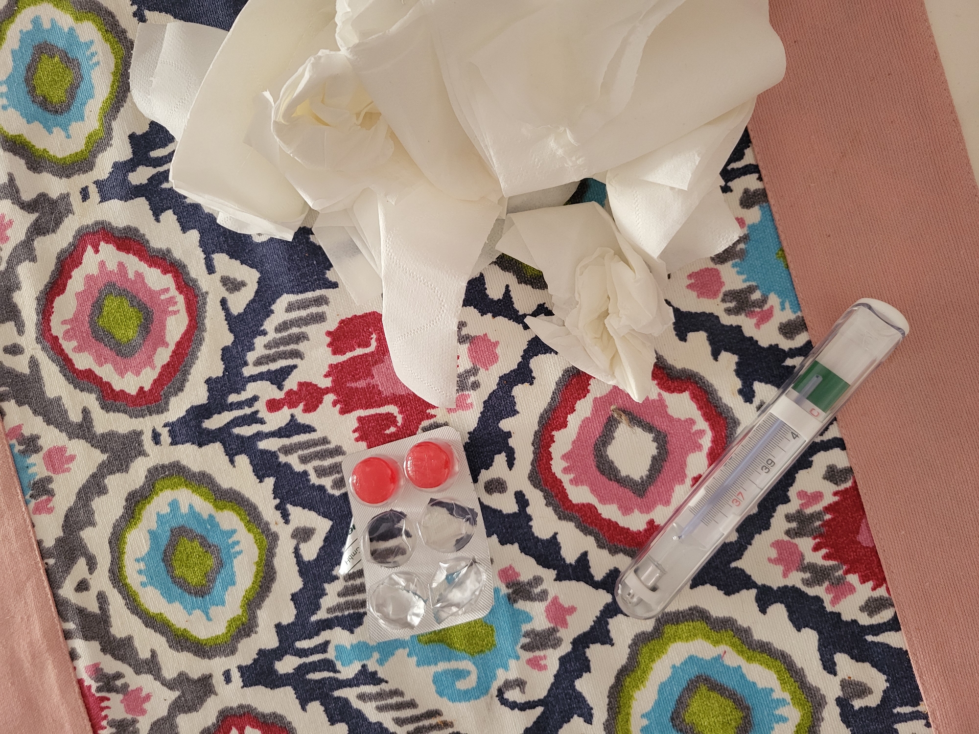 The image shows a still life composition on a patterned fabric surface. The fabric features colorful, circular motifs of blue, red, and pink against a white background with navy blue accents. Scattered on the fabric are crumpled white tissues, a thermometer indicating a temperature around 38°C, and a blister pack of red lozenges. A section of pink fabric is visible in the upper right corner of the image.
The photograph does not depict any people, actions, or interactions, and there are no elements indicating that the location is in Budva, Montenegro.