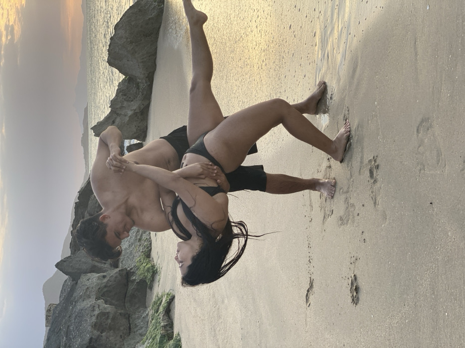 On a sandy beach in Juan Griego, Venezuela, a male and a female are engaged in a dance pose at sunset. The male, bare-chested and wearing dark shorts, stands upright, supporting the female. His left arm is around her waist, and his right hand grasps her left hand. The female, wearing a dark two-piece swimsuit, is leaned backward, with her body horizontally inclined. Her left leg is extended straight backward and upward, while her right leg is bent with her foot on the sand. Her right arm is positioned around the male's neck/shoulder. Both individuals are barefoot. Behind them, large, dark rocks, some covered with green growth, line the beach. The background features the ocean with a visible horizon and distant landmasses under a sky showing soft orange and yellow hues, indicative of a sunset. The sand in the foreground displays footprints and appears slightly damp near the water's edge.