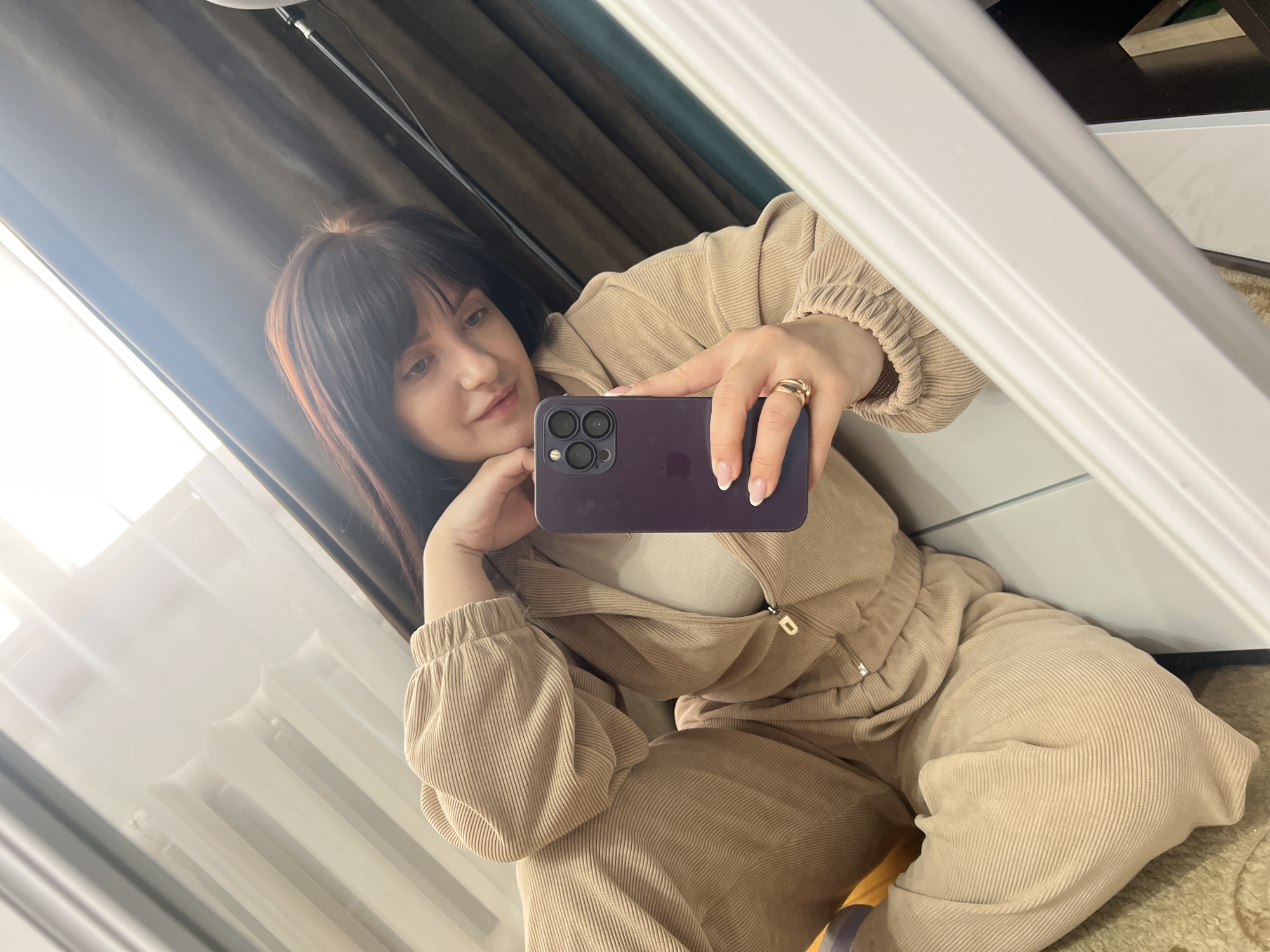 A woman is taking a selfie in a mirror. She is wearing a tan corduroy tracksuit. She is holding a purple iPhone in her right hand. She has dark hair and is wearing a gold ring on her right ring finger. The background includes curtains, a window, and a white wall. The location is Марковский, Russia.