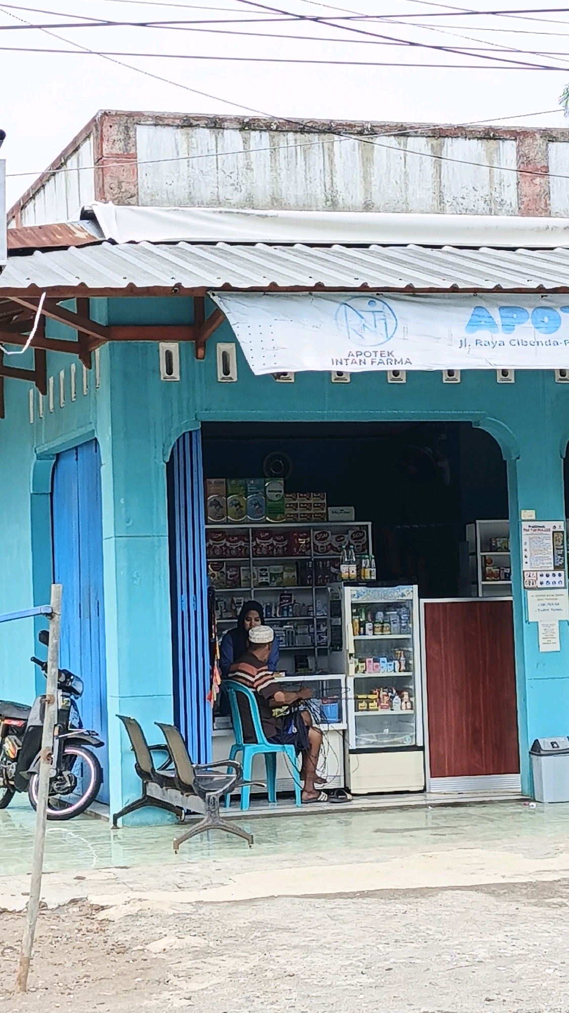 The image captures a storefront in Cibenda, Indonesia, with a blue facade and a corrugated metal awning. A white banner across the awning reads "APOTEK INTAN FARMA" and "Jl. Raya Cibenda-P." Two individuals are visible inside the open storefront. One, a person wearing a headscarf and a striped shirt, appears to be an employee or the owner, interacting with a customer seated on a blue chair. The customer, a man wearing a white skullcap and a striped shirt, is looking down at something in his hands. Shelves stocked with various products, including what appear to be medicines and bottled drinks, are visible behind them. A glass-fronted refrigerator displays more items. In front of the store, two empty metal chairs and a parked motorcycle are on the tiled walkway. The street in front of the establishment is paved with concrete. The overall scene depicts a small business in a street setting.
