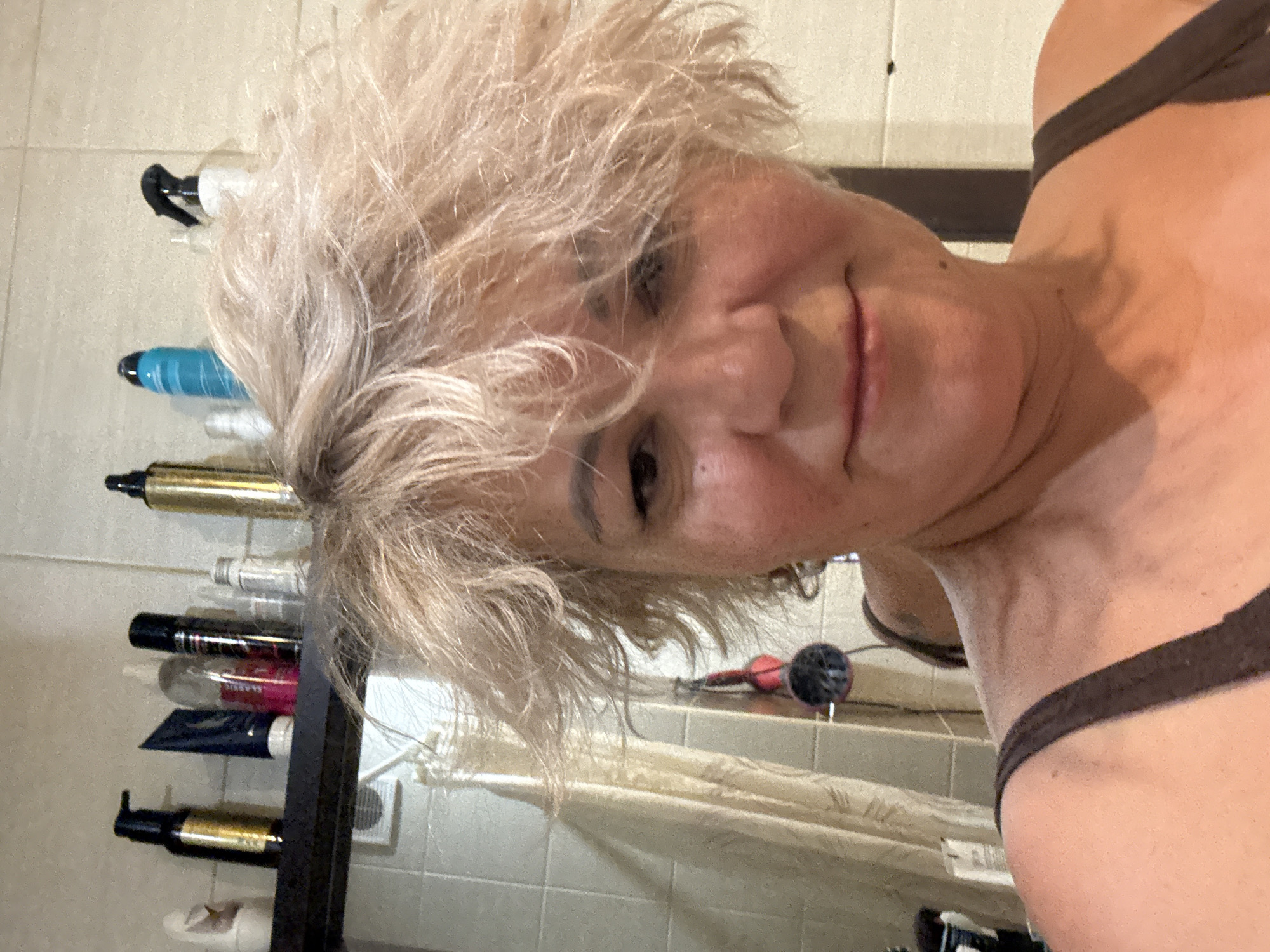 A woman with blonde, tousled hair takes a selfie in a bathroom. She is wearing a dark brown tank top. Behind her, on a shelf, are several bottles and containers of toiletries. The walls are tiled with a light-colored tile. A shower curtain is visible to the left. The lighting suggests an indoor setting. The image does not provide any information about the city of Kazan, Russia.