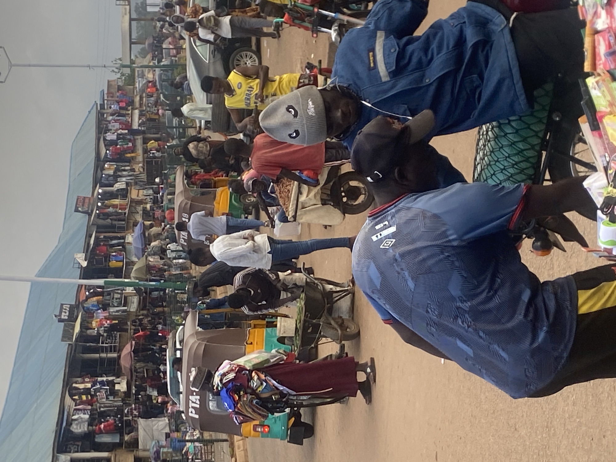In the city of Bukuru, Nigeria, a bustling outdoor market scene unfolds. The foreground is dominated by several men, two of whom are seated on a motorcycle. One man, wearing a blue jacket and a gray beanie with cartoon eyes, appears to be interacting with the man in front of him. The second man, facing away from the camera, wears a blue shirt with a patterned design and black pants with yellow accents. In the mid-ground, a variety of individuals are engaged in market activities. A man in a dark shirt and light pants is using a wheelbarrow filled with green items, possibly produce. Nearby, a person in a maroon robe carries a large bundle of folded clothes. A three-wheeled vehicle, possibly a taxi or delivery van, is parked behind them. Further into the background, a row of market stalls displays a wide assortment of clothing items. Cars and other vehicles are visible on the road, indicating a busy commercial area. The overall atmosphere suggests a vibrant and active marketplace.