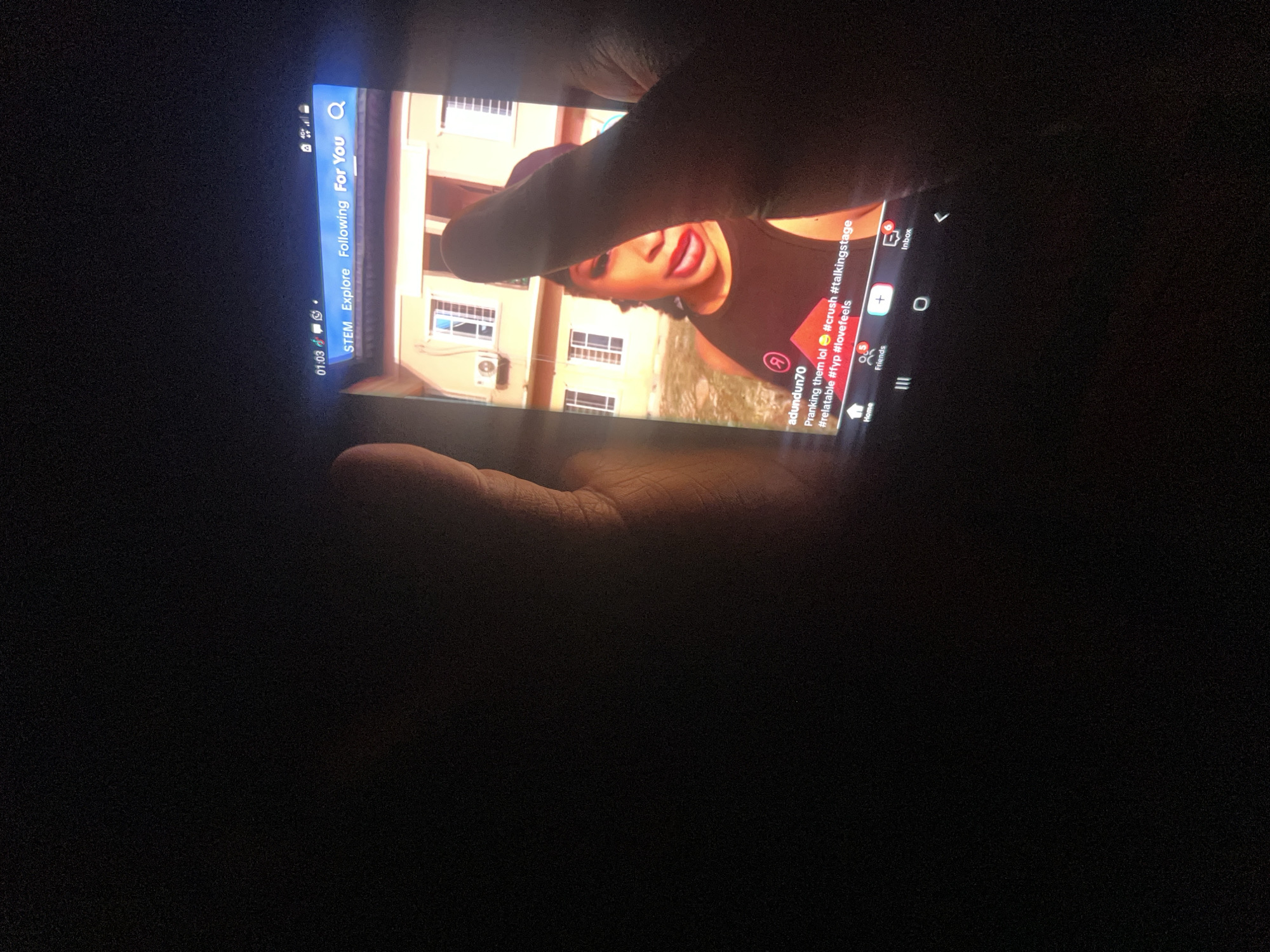 A vertical close-up shot shows a smartphone being held by two hands in a dark environment. The phone screen is illuminated and displays a TikTok video. The video features a woman with red lipstick and a black top. The background of the video shows a building with windows and an air conditioning unit. Text overlay on the video reads "adundun70 Pranking them lol #crush #talkingstage #relatable #fyp #lovefeels". A vertical bar on the right side of the screen displays "STEM Explore Following For You" and a search icon. The timestamp on the video reads "01:03". The overall scene suggests a casual setting, possibly in Nkpor, Nigeria, where the user is engaged with social media content in low light conditions.