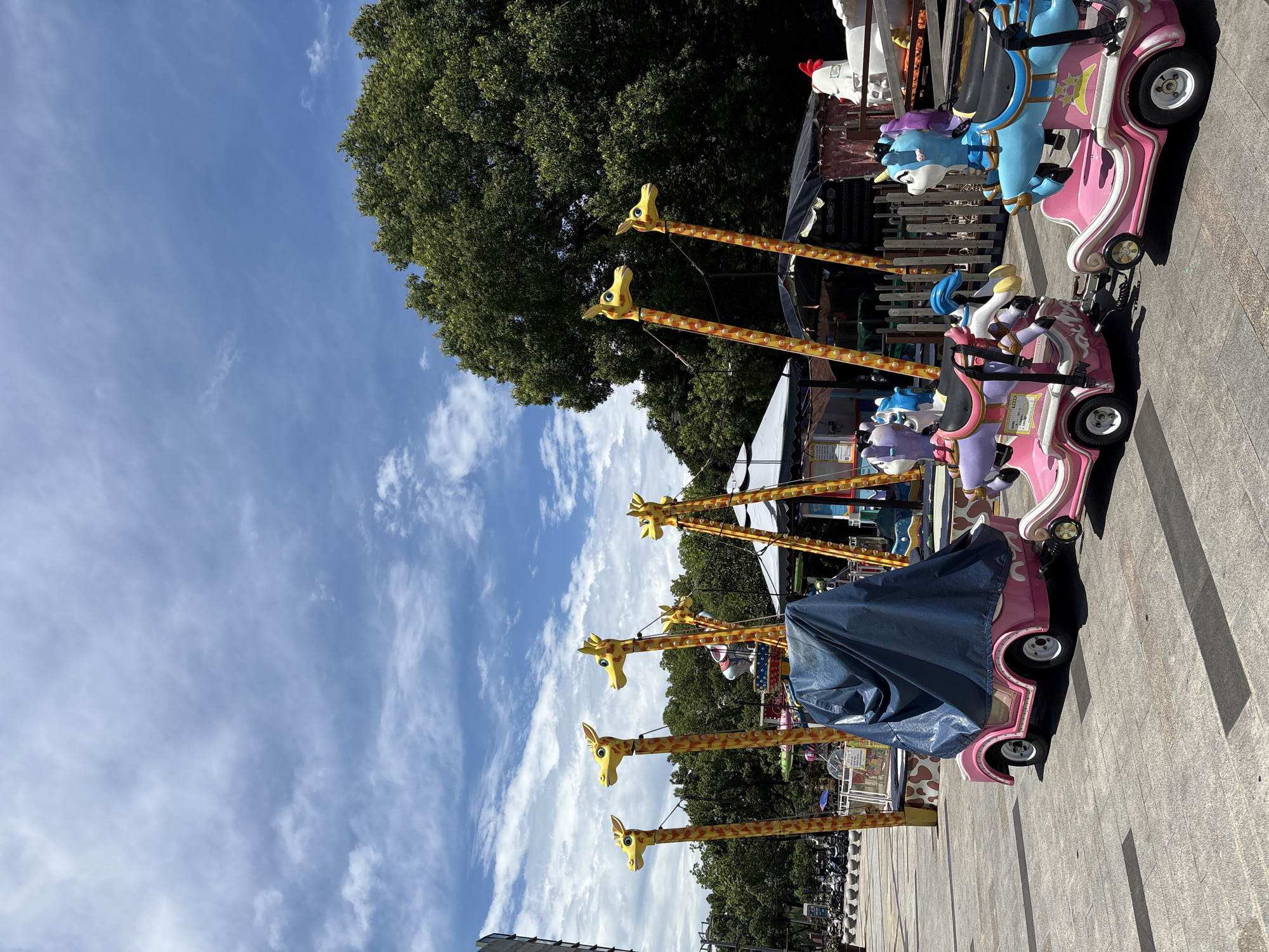 The image depicts a sunny outdoor scene in Minhang District, China, featuring a children's amusement ride. The ride consists of several colorful, cartoon-like cars, some of which are adorned with unicorn figures. The cars are connected and appear to be waiting to be used. In the background, tall, yellow, giraffe-shaped poles with orange spots rise towards a partly cloudy blue sky. A large, lush green tree dominates the right side of the frame. The ground is paved with square tiles, and a few parked motorcycles are visible on the left, suggesting the location is a public area. One of the cars is covered with a dark blue tarp.