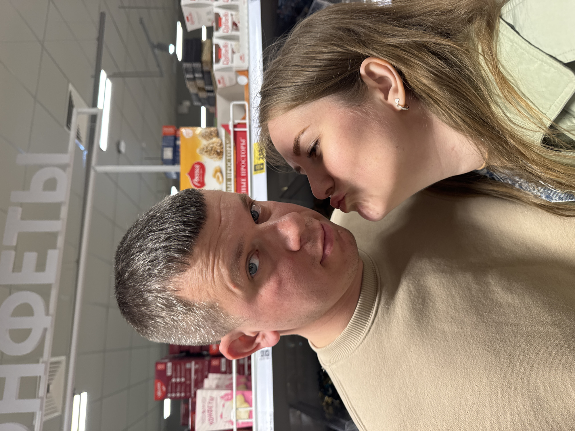 A man and a woman are posing close together in a store aisle. The woman appears to be playfully trying to kiss the man's cheek. They are surrounded by shelves stocked with various packaged goods. The location is Горный, Russia.