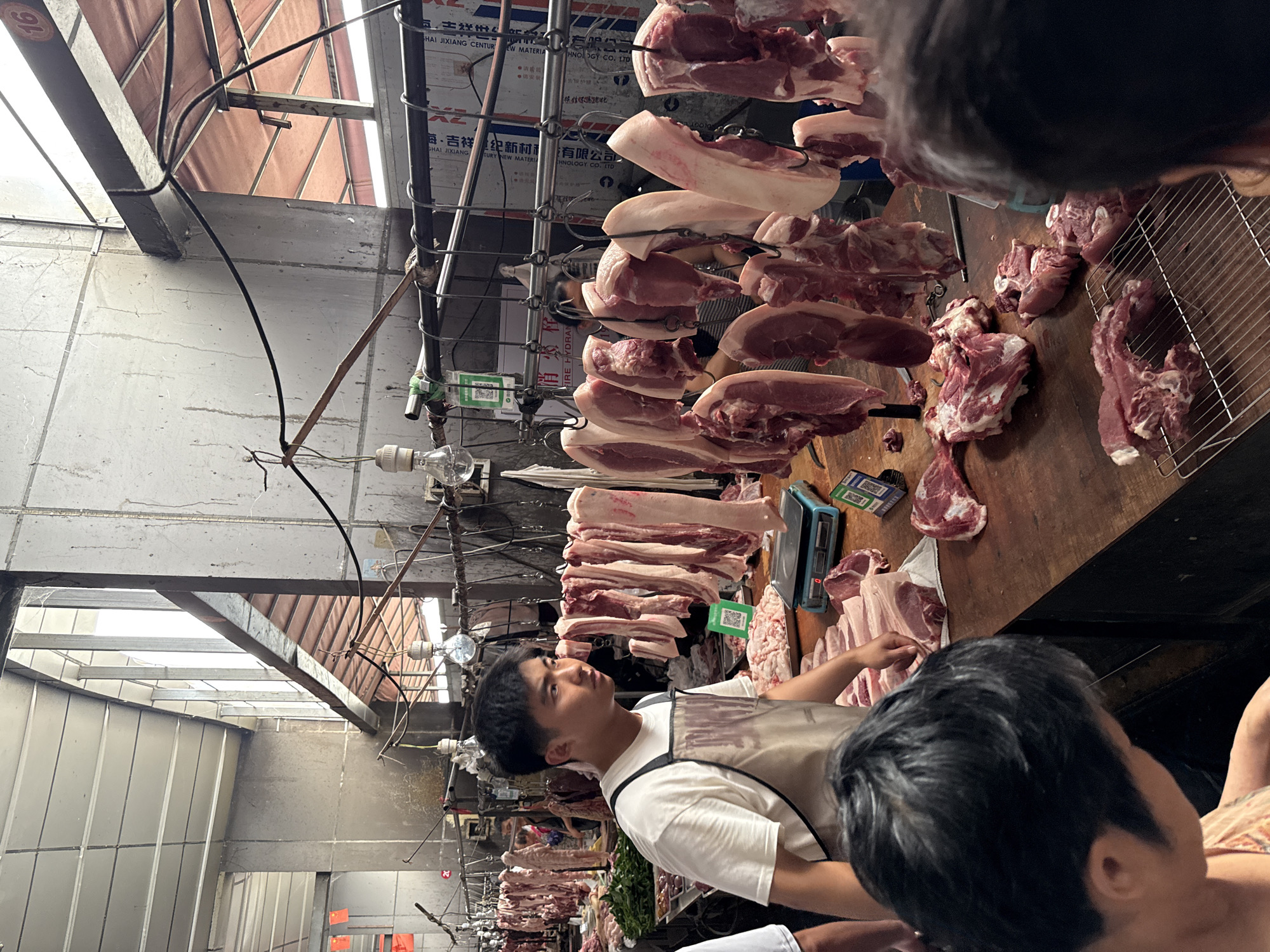 This image captures an active indoor meat market stall in Jiangjin, China. A young East Asian male, wearing a white t-shirt and a dark apron, stands behind a wooden counter, looking right. His stall prominently displays numerous cuts of raw pork; large sections with skin and fat hang from metal hooks on overhead rails, while various smaller cuts are arranged on the counter. Two digital scales, one blue and one black, are visible on the counter, alongside piles of fresh meat, including bone-in pieces on a metal grate. Multiple QR code stickers, likely for mobile payments, are affixed to the counter and hanging fixtures.
Several customers are partially visible in the foreground and background, indicating an active commercial environment. The market features concrete walls, some appearing weathered, and a tiled ceiling. Exposed electrical wiring and bare light bulbs provide illumination, supplemented by natural light entering from an upper opening that reveals a reddish awning structure outside. In the background, a metal panel is visible with text including "HAI JIANGJIANG CENTURY NEW MATERIAL TECHNOLOGY CO. LTD" and "FIRE HYDRANT," confirming the location in Jiangjin. A red sign with the number "16" is in the upper right, and another with "20" is further left, possibly indicating stall numbers.