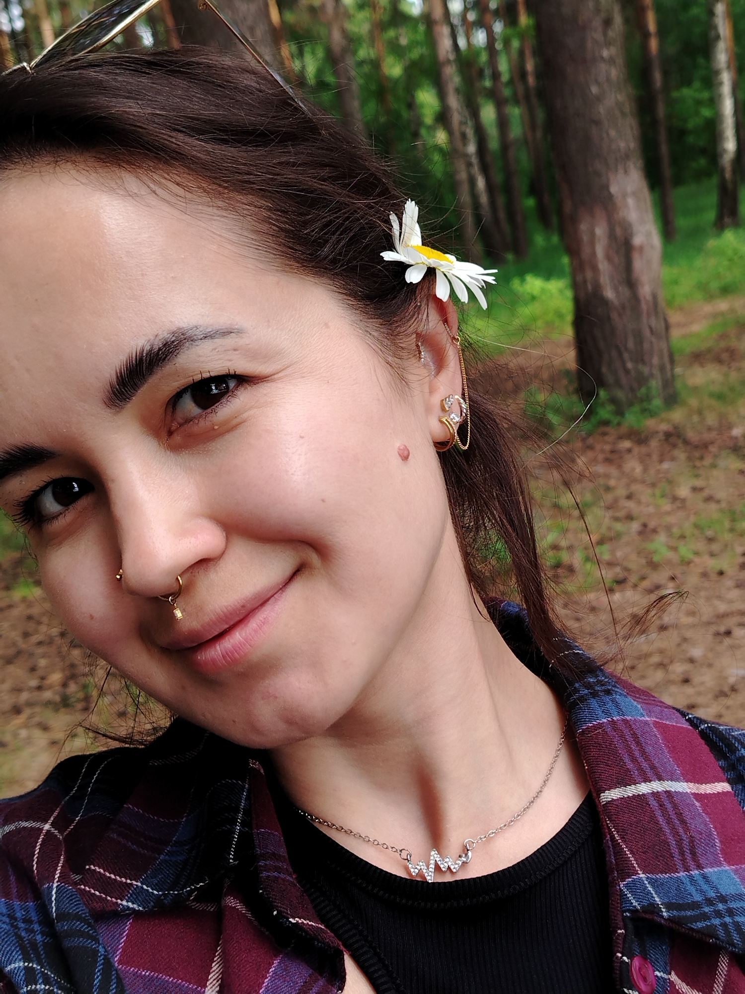 A close-up portrait captures a young woman smiling warmly at the camera in a natural, wooded setting identified as Chaikovsky, Russia. She has dark hair and dark eyes, with a joyful and relaxed expression. A white and yellow daisy is playfully tucked behind her right ear, and the top of sunglasses rests on her head, partially visible.
Her facial features include a small golden stud in her left nostril and a golden septum ring featuring a dangling cube. Her right ear is adorned with multiple intricate gold earrings, including a cuff, studs, and a delicate chain. A small, light brown mole is visible on her right cheek.
She wears a plaid button-up shirt in shades of maroon, blue, and black, layered over a black ribbed top. Around her neck is a silver chain necklace with a sparkling, crystal-embellished design resembling a heart-rate or ECG line. The background is softly blurred, showing tall tree trunks and green foliage, indicating a forest or park environment under what appears to be pleasant daytime lighting.