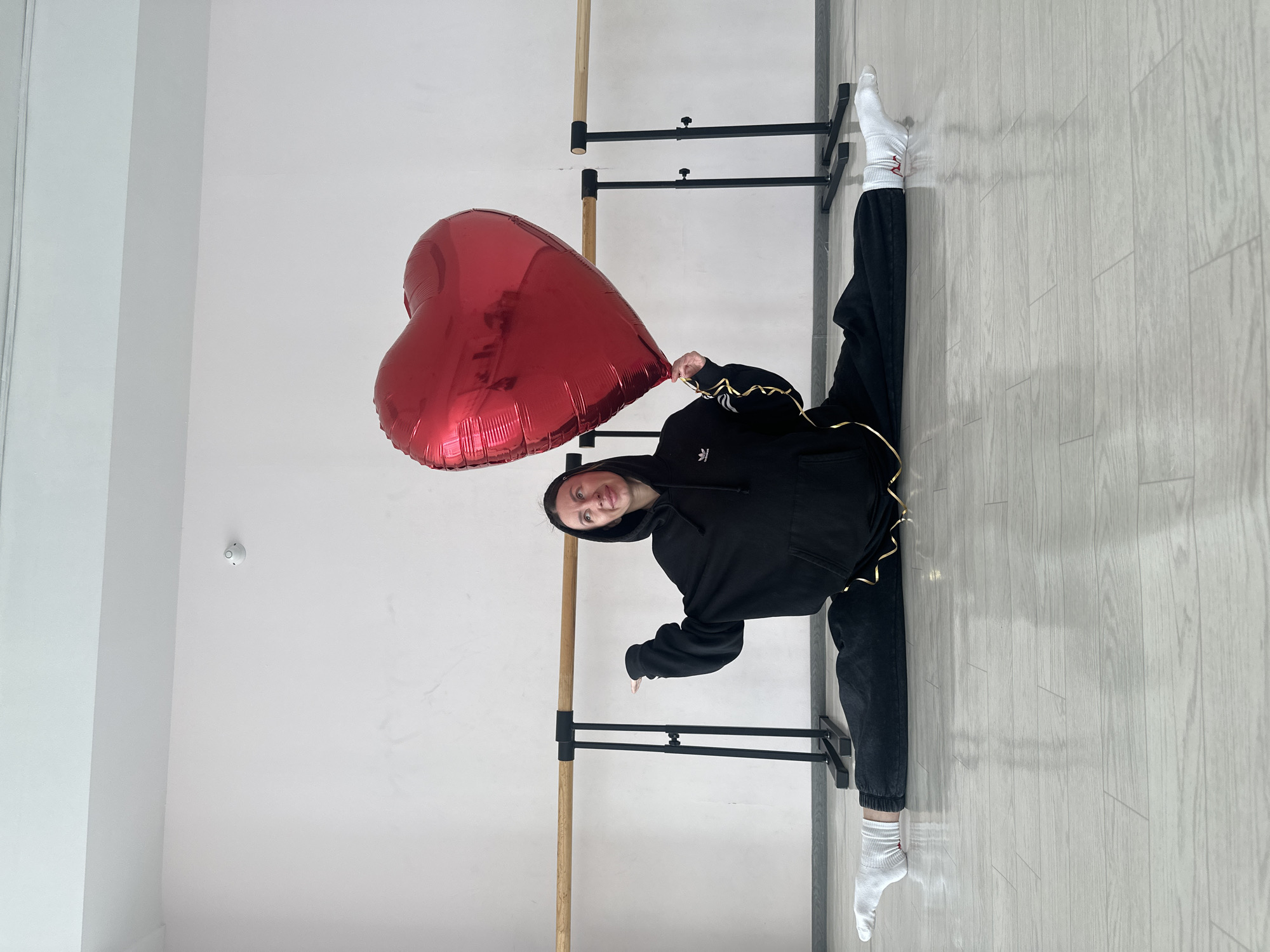 A young woman in a black hooded sweatshirt and black pants is performing a split in a room with wooden floors. She is holding a large red heart-shaped balloon. Her legs are extended, with one foot against a ballet barre. She's wearing white socks. The room appears to be a dance studio with a barre attached to the wall. The walls are white. The image was taken at an angle, making the scene appear sideways. There are two-tiered ballet barres in the background. The image location is Votkinsk, Russia.