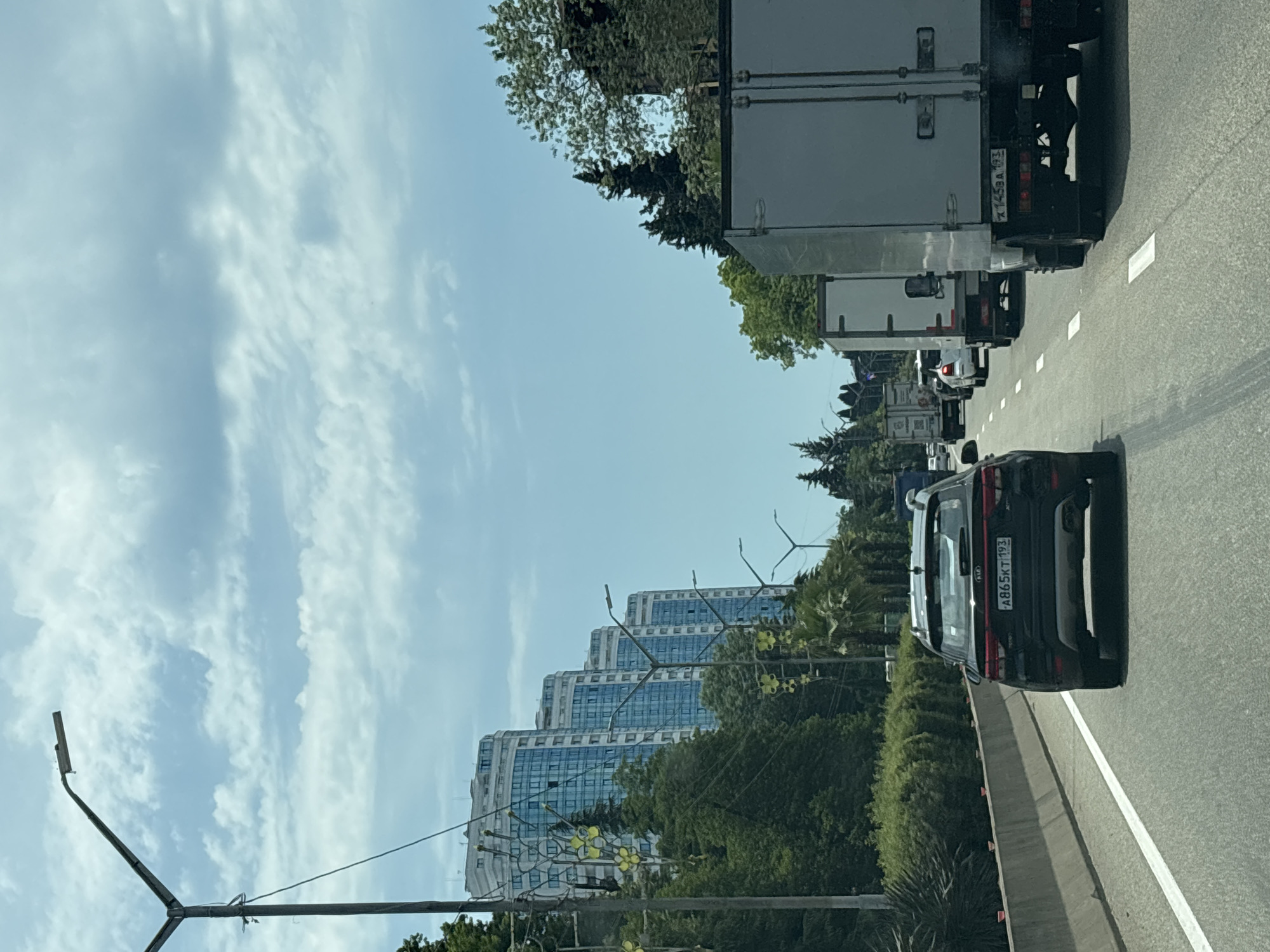 The image depicts a sunny day in Sochi, Russia, likely on a highway. In the foreground, a black Kia SUV is driving on a multi-lane road. To the right of the SUV, a large white truck is visible. The road is lined with a concrete barrier on the left, beyond which are lush green trees and bushes. In the background, several tall, modern apartment buildings with blue-tinted windows are prominent, suggesting an urban or resort area. Streetlights are also visible along the roadside. The sky is blue with scattered white clouds. The overall scene is of moderate traffic on a clear day in a coastal city.