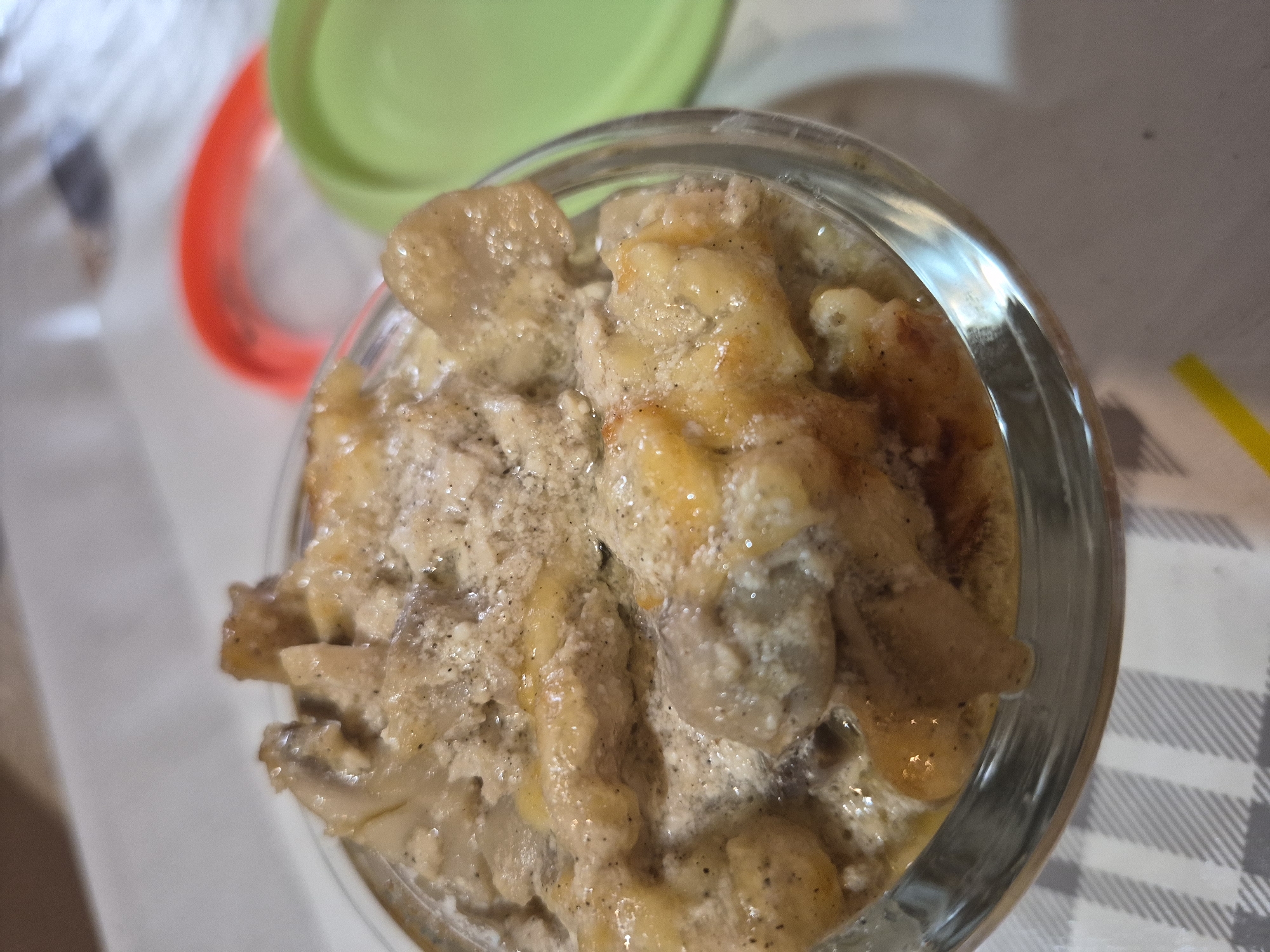 A close-up, high-angle shot shows a clear glass bowl filled with a creamy, light brown stew. The stew contains pieces of meat, possibly chicken or pork, and some yellow ingredients, possibly potatoes or corn. The dish is seasoned with black pepper, visible as small dark specks throughout the creamy sauce. The bowl sits on a grey and white checkered tablecloth. In the background, a red lid is visible to the left and a green lid is visible to the right, both out of focus. The lighting is soft, with some reflections on the glass bowl. The scene appears to be indoors, possibly in a home kitchen or dining area in Yerevan, Armenia.