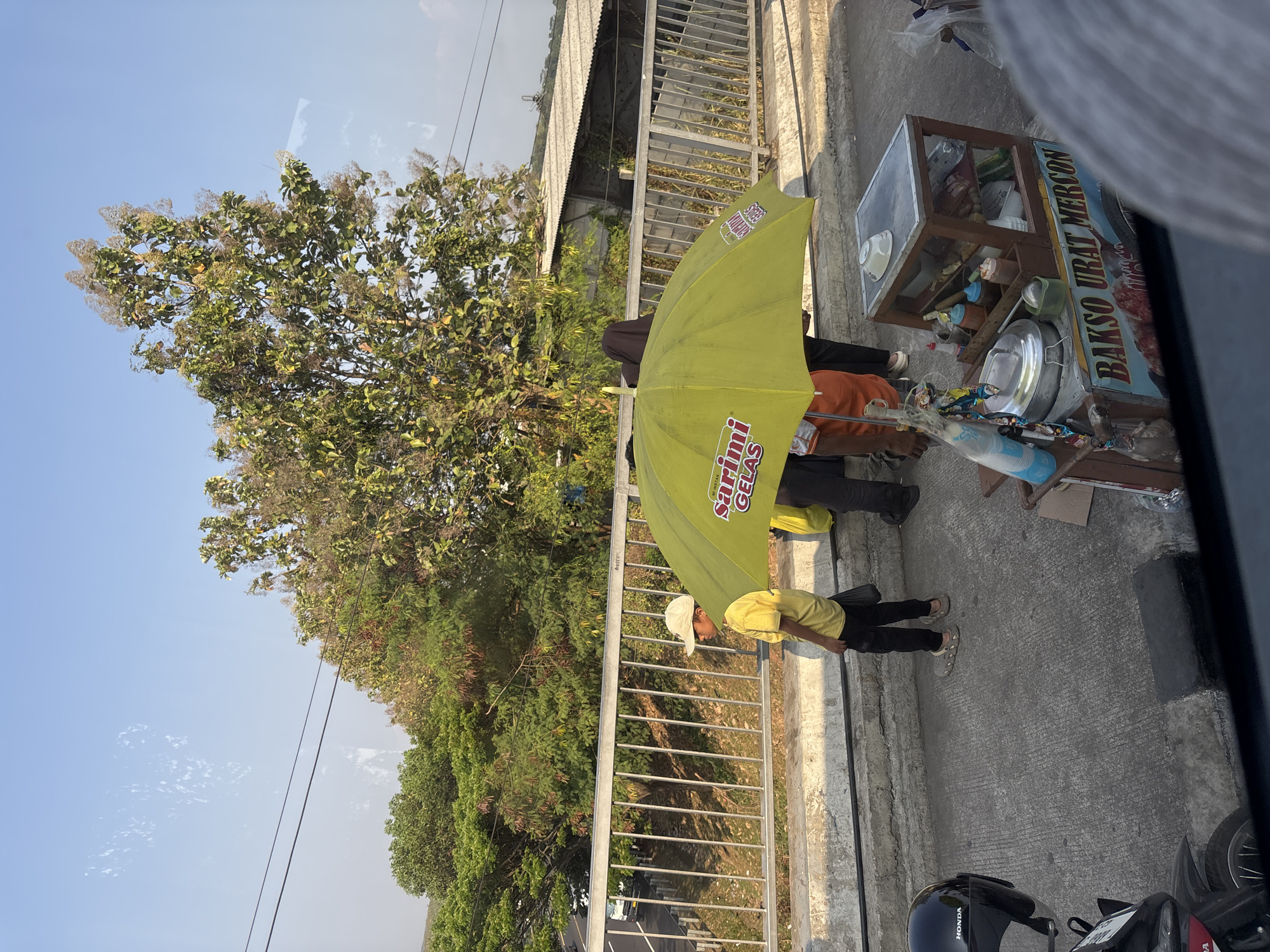 The image shows a street scene in Cikampek, Indonesia, on a clear day. A young boy wearing a yellow shirt and a white cap stands under a bright yellow umbrella. Next to him is a food cart selling "Bakso Urat Mercon". The cart has glass displays filled with ingredients and condiments. Behind the boy and the cart, a person wearing a dark headscarf and dark clothing is visible. In the background, there is a metal fence, lush green trees, and buildings with corrugated roofs. A motorcycle helmet rests on the ground to the left of the frame, and a portion of a motorcycle is visible at the bottom left. The overall scene suggests a typical day in a bustling Indonesian city.