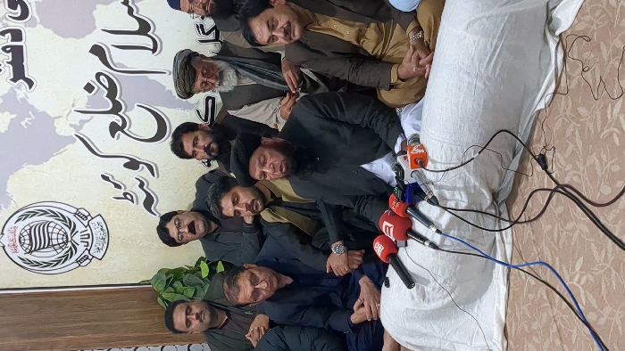 A group of men, seemingly politicians or community leaders, are gathered together. Microphones from different news agencies are positioned in front, suggesting a press conference or important announcement. The backdrop features a logo and writing in a non-English language, possibly Urdu or Arabic. The overall setting appears to be indoors, with a patterned wall and some greenery visible. The mood is somber, and some of the men seem contemplative or tired.