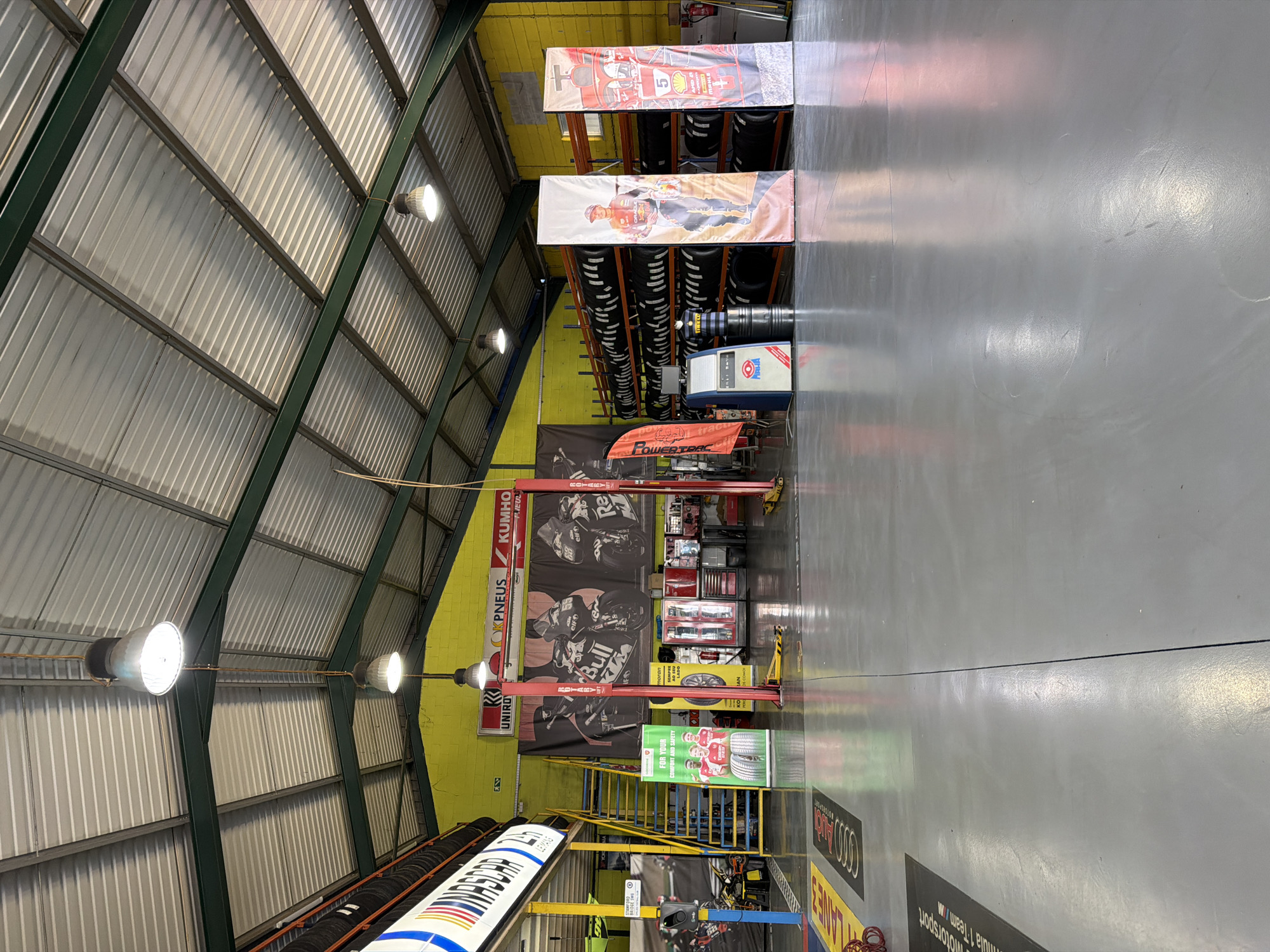 The image depicts the interior of a tire shop in Vila Real, Portugal. The space features a high, corrugated metal ceiling with exposed green beams and hanging industrial lights. Walls are painted yellow and gray. The shop houses shelves stacked with tires, banners displaying racing themes, and equipment such as a tire changer and a car lift. There are no people visible. Banners for brands like Kumho, Uniro, and Red Bull are seen. Signage includes motorsport and Le Mans references.
