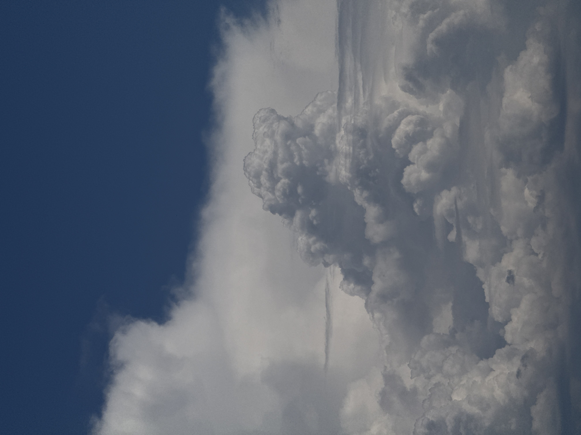 A vertical, medium shot captures a dramatic sky filled with a vast expanse of textured cumulus clouds against a deep blue upper atmosphere. The clouds are a mix of bright white and deep grey, with some appearing soft and fluffy while others are dense and ominous, suggesting a stormy or turbulent weather system. The bottom portion of the image shows darker, heavier cloud formations, giving way to lighter, more illuminated clouds higher up. The overall impression is one of immense scale and natural power, emphasizing the dynamic beauty of the sky. The scene is devoid of any man-made structures or living beings, focusing solely on the atmospheric phenomenon. The location is stated as Unknown, Unknown.