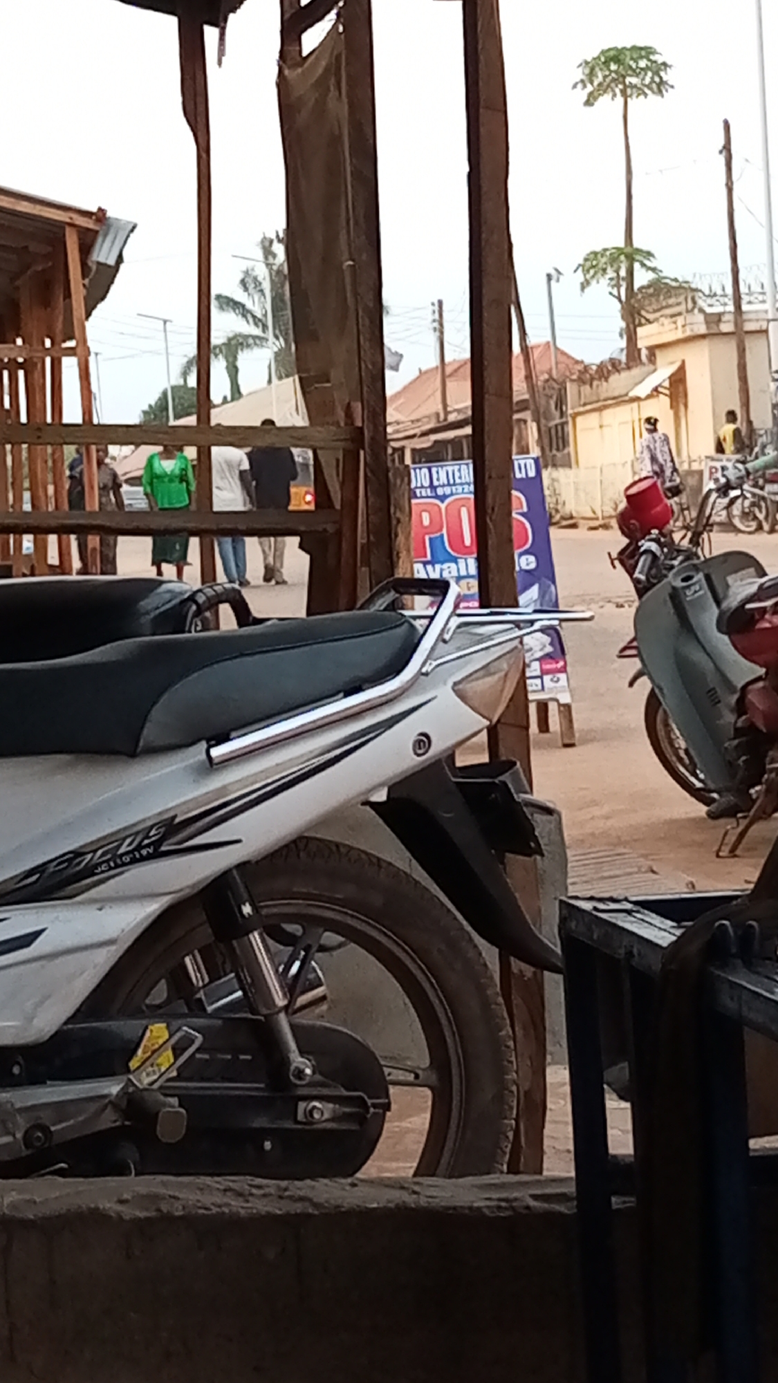 The image, taken in Sabon Tasha, Nigeria, shows a street scene viewed from a partially obstructed vantage point. Dominating the foreground are parked motorcycles. A white motorcycle with black accents, labeled "FOCUS," is prominently positioned on the left side. The wheel and rear portion of the motorcycle are in clear focus. To the right, a portion of another motorcycle with a red gas can is visible.
In the background, there is a view of a street. Several people are present, some walking along the sidewalk. A sign for "JIO ENTERPRISE LTD" is visible. The architecture includes buildings with corrugated metal roofs and utility poles. A few sparse trees are also visible. The overall scene is one of a typical urban street environment in a Nigerian city.