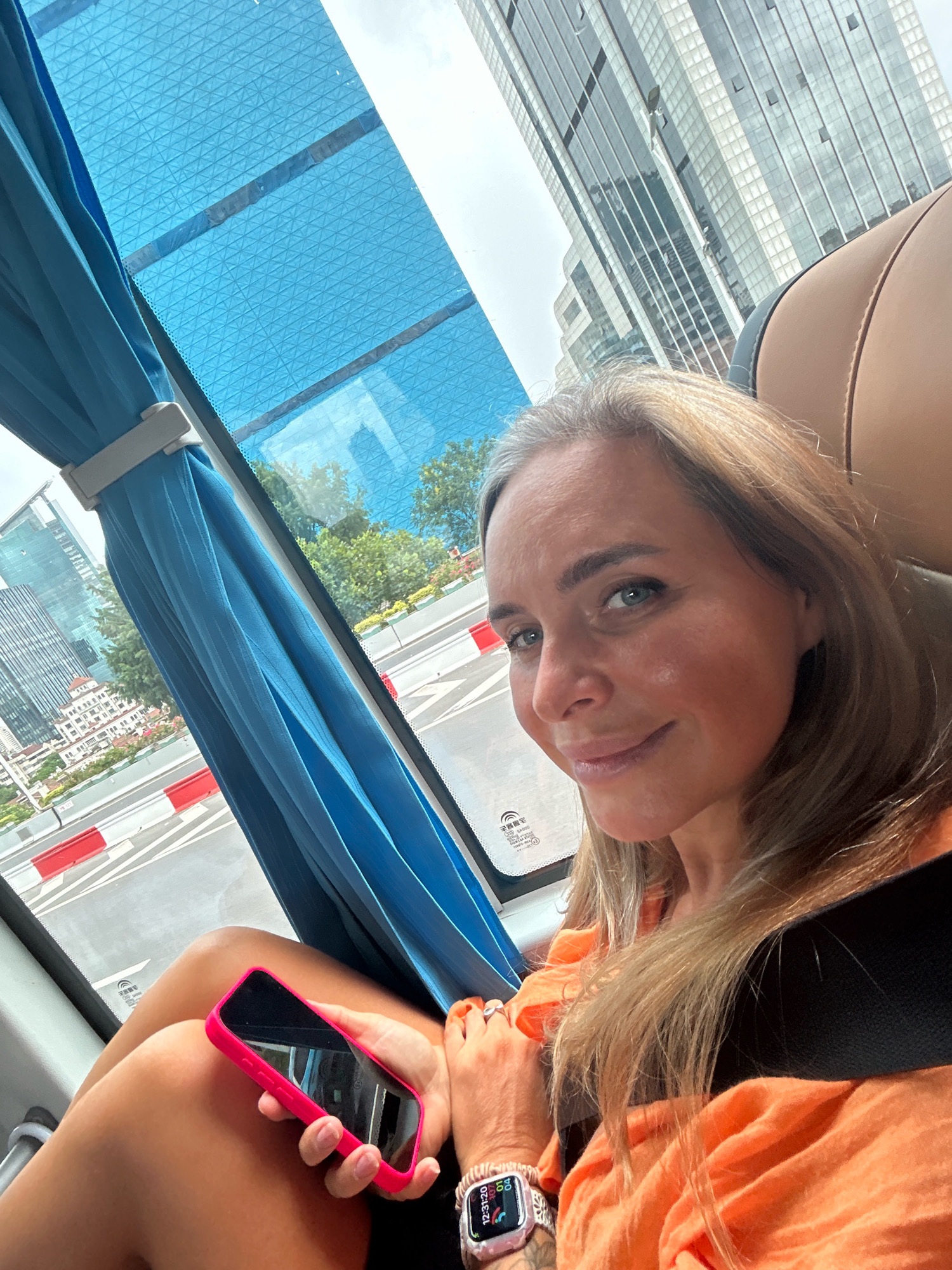 A woman is sitting in a vehicle, likely a bus, in Jing'an District, China, holding a pink smartphone. She has blonde hair and is wearing an orange shirt. A seatbelt is visible across her chest. Outside the window, modern skyscrapers and trees are visible, indicating an urban environment. The window has a blue sunshade.