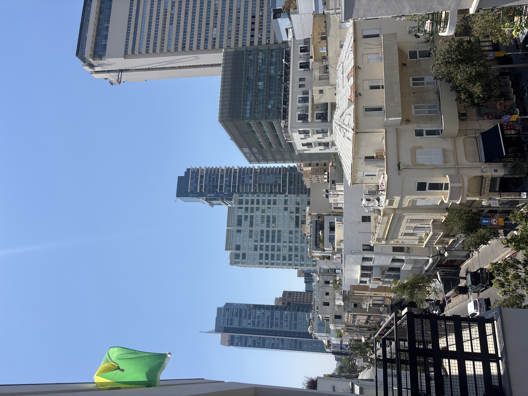 This image depicts a city street scene in Tel Aviv, Israel, characterized by a mix of modern and older architecture under a clear blue sky. A green and yellow flag is visible in the upper left corner, attached to a building. In the background, several tall, modern skyscrapers with glass facades dominate the skyline, including a prominent white skyscraper on the right. In the middle ground, there are more contemporary office buildings with horizontal louvers.
Closer to the street level, a row of older, low-rise buildings with light-colored facades and balconies lines the street. These buildings appear to house shops and cafes, with some outdoor seating visible. A black awning is extended over a storefront. The street itself is paved and shows signs of urban activity. Two cars, a white sedan and a dark pickup truck, are parked on the street, and a few individuals are walking. Traffic signals and street signs, including one with the number "50", are present. Trees are scattered along the street and on balconies, adding greenery to the urban landscape. The vantage point suggests the photo was taken from a higher level, possibly a balcony or window of a building overlooking the street.