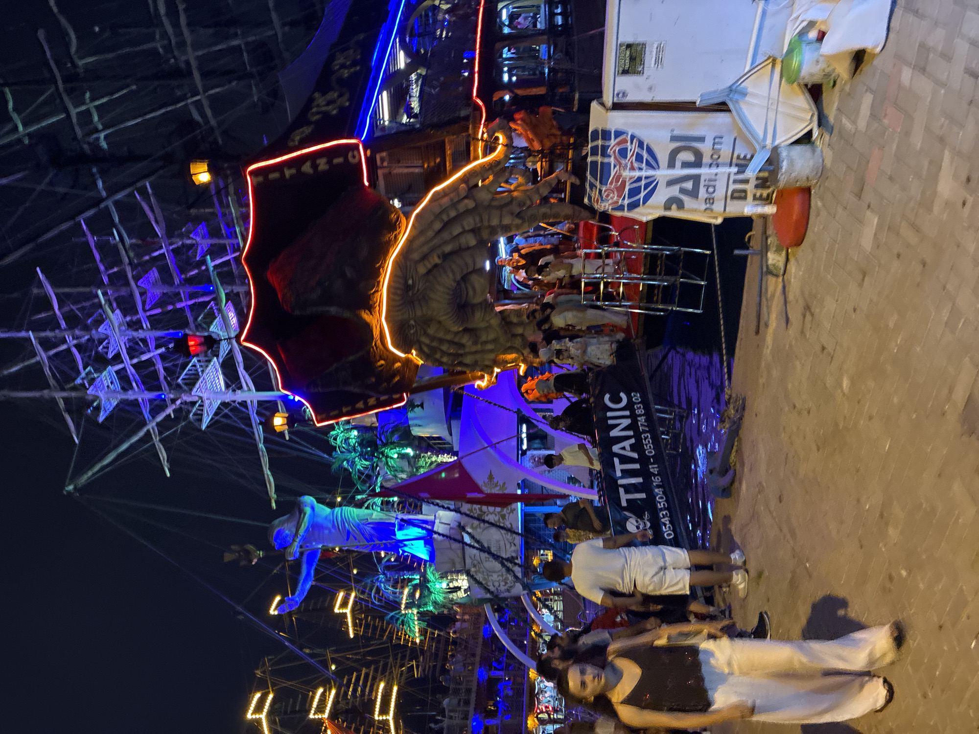 This media file captures a lively scene at night, likely a waterfront area, possibly a tourist spot in Alanya, Turkey. The primary focus is on a decorative, illuminated boat structure resembling a pirate ship, adorned with vibrant neon lights in blue and red. A large, sculpted hand, also outlined in glowing orange neon, is a prominent feature of the boat's decoration, creating a dramatic and eye-catching effect.
Several people are visible in the foreground and around the boat, suggesting an event or gathering is taking place. Some are standing, while others are seated. Their clothing appears casual, and the atmosphere seems festive, enhanced by the colorful lighting and the overall visual spectacle. The ground is paved, indicating an urban or developed area.
A large banner on the boat displays the word "TITANIC" in bold lettering, along with a phone number: "0543 504 16 41 - 0553 774 83 02". Another banner shows a logo with the word "PADI.com", suggesting a connection to scuba diving or water activities. The presence of palm tree decorations further reinforces a tropical or coastal setting. The time of day is clearly night, as evidenced by the darkness and the prominence of the artificial lights. The weather appears to be clear, allowing for outdoor activities.