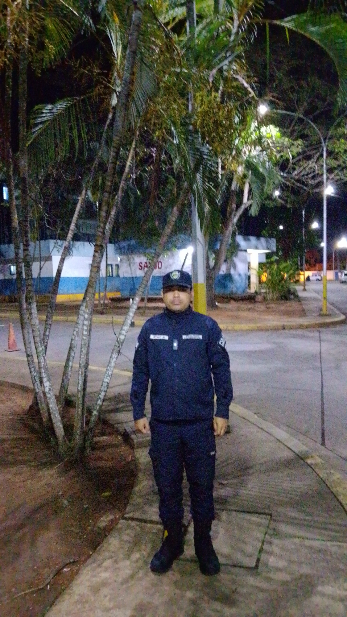 An adult man, likely a security guard or officer, stands centrally in a nighttime outdoor setting in Tinaquillo, Venezuela. He is wearing a dark navy blue uniform, consisting of a long-sleeved shirt with a collar, matching trousers, and a dark cap with an emblem that is not fully legible. The name "WEBBCO JR" is visible on his left chest. He has a neutral expression and is facing the camera directly.
The background features several tall palm trees with fronds extending into the dark sky, some illuminated by artificial light. Behind the trees, a building with white and blue sections is visible, and the partial text "SAUD" can be seen on its facade, suggesting a medical or health-related facility ("SALUD"). Streetlights are scattered throughout the background, casting light on the paved road and sidewalk where the man stands. An orange traffic cone is present to the left of the man, near the base of the palm trees. The scene is entirely lit by artificial sources, confirming it is nighttime.