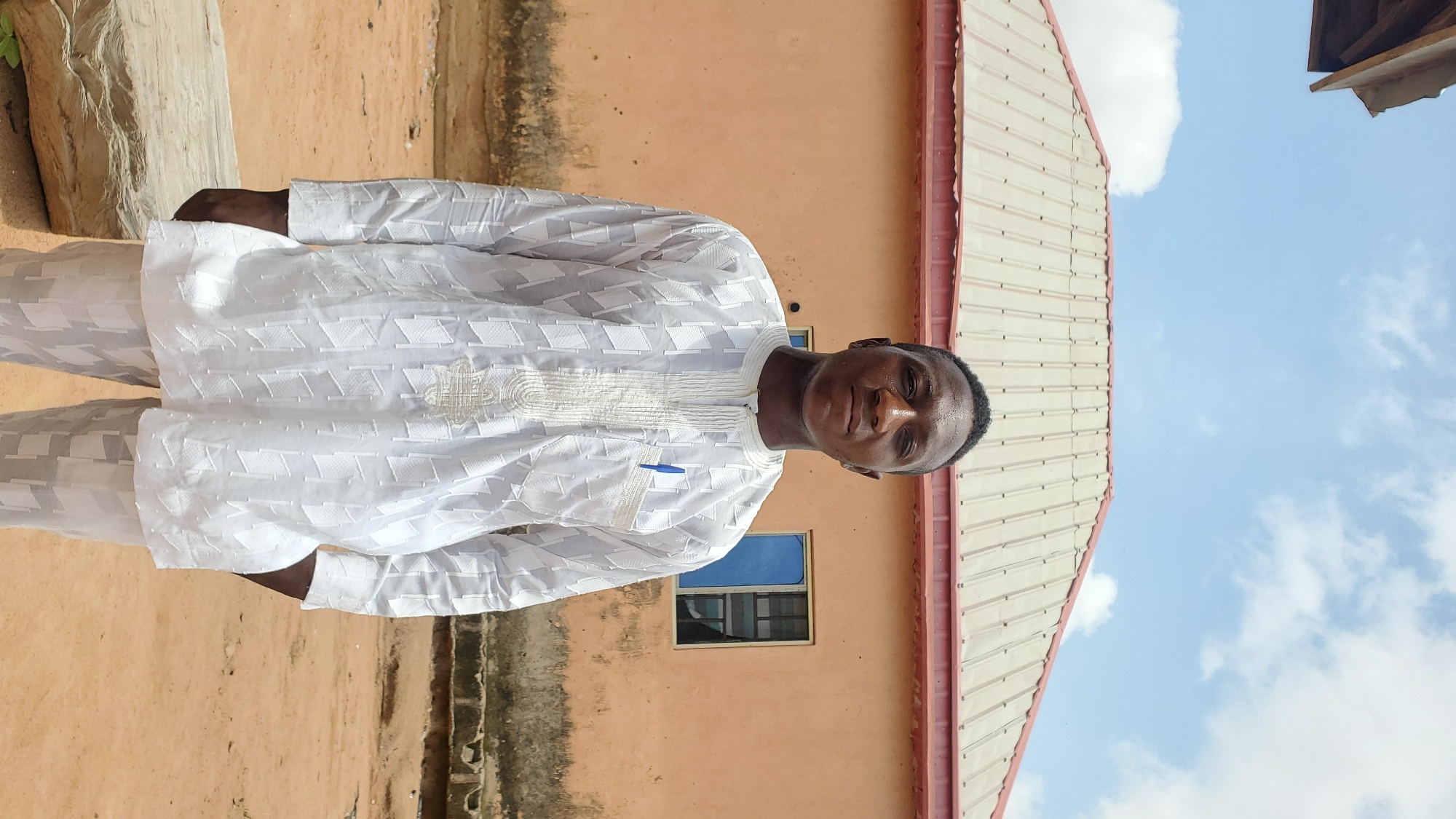 A young Black male stands outdoors in front of a two-story building, with a light beige facade and a pale cream-colored corrugated metal roof. He is wearing a white traditional African outfit consisting of a long-sleeved tunic with intricate white embroidery around the neckline and chest, and matching white trousers. A blue pen is tucked into the left breast pocket of his tunic. His hands are in his pockets, and he is looking directly at the camera. The ground in front of him is sandy and uneven, with a large, weathered wooden log on the left side of the frame. Behind the man, a blue-tinted window is visible on the building. The sky above is a bright blue with scattered white clouds. The building and the sandy ground suggest a location in Jalingo, Nigeria.