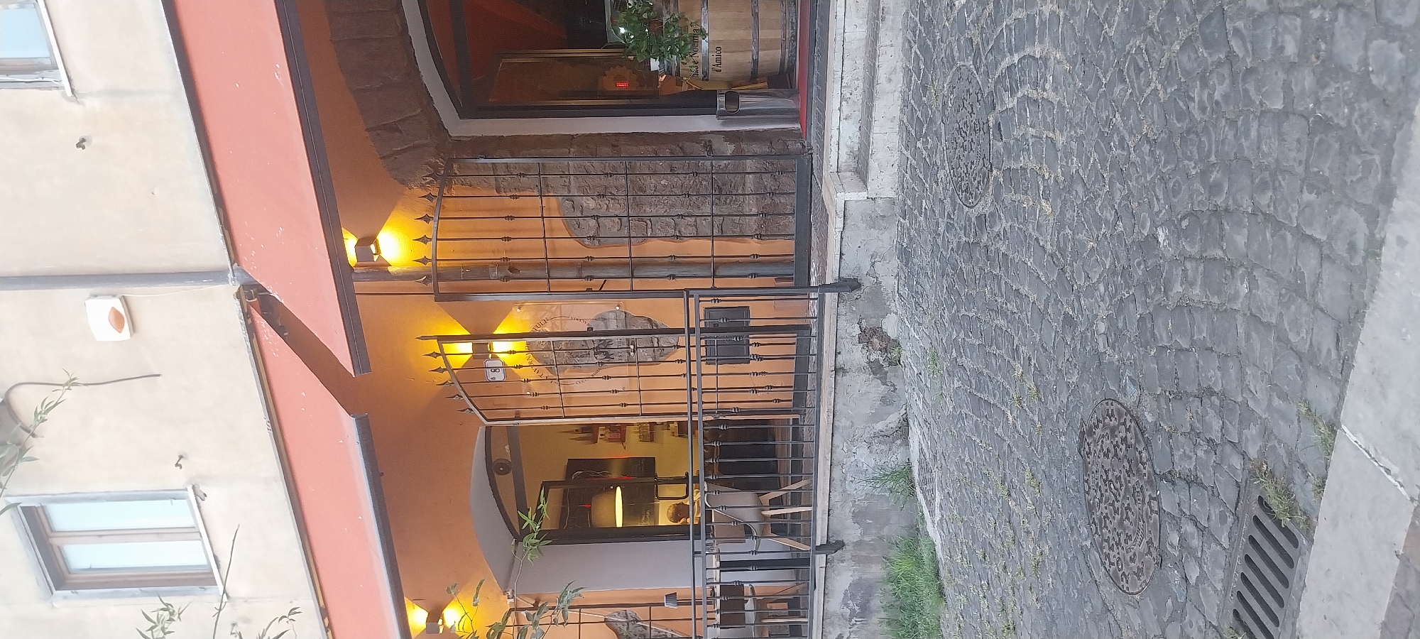 The image shows a storefront in Ardea, Italy, with an outdoor seating area. The storefront features an orange facade with red awnings and is lit by exterior lights. A black metal gate is open, revealing the entrance to the establishment. Inside, a person is visible sitting at a table. In front of the building, a cobblestone street is visible, with a manhole cover and a drain. To the right of the entrance, a wooden barrel is placed outside. The building appears to be old, with visible stone elements.