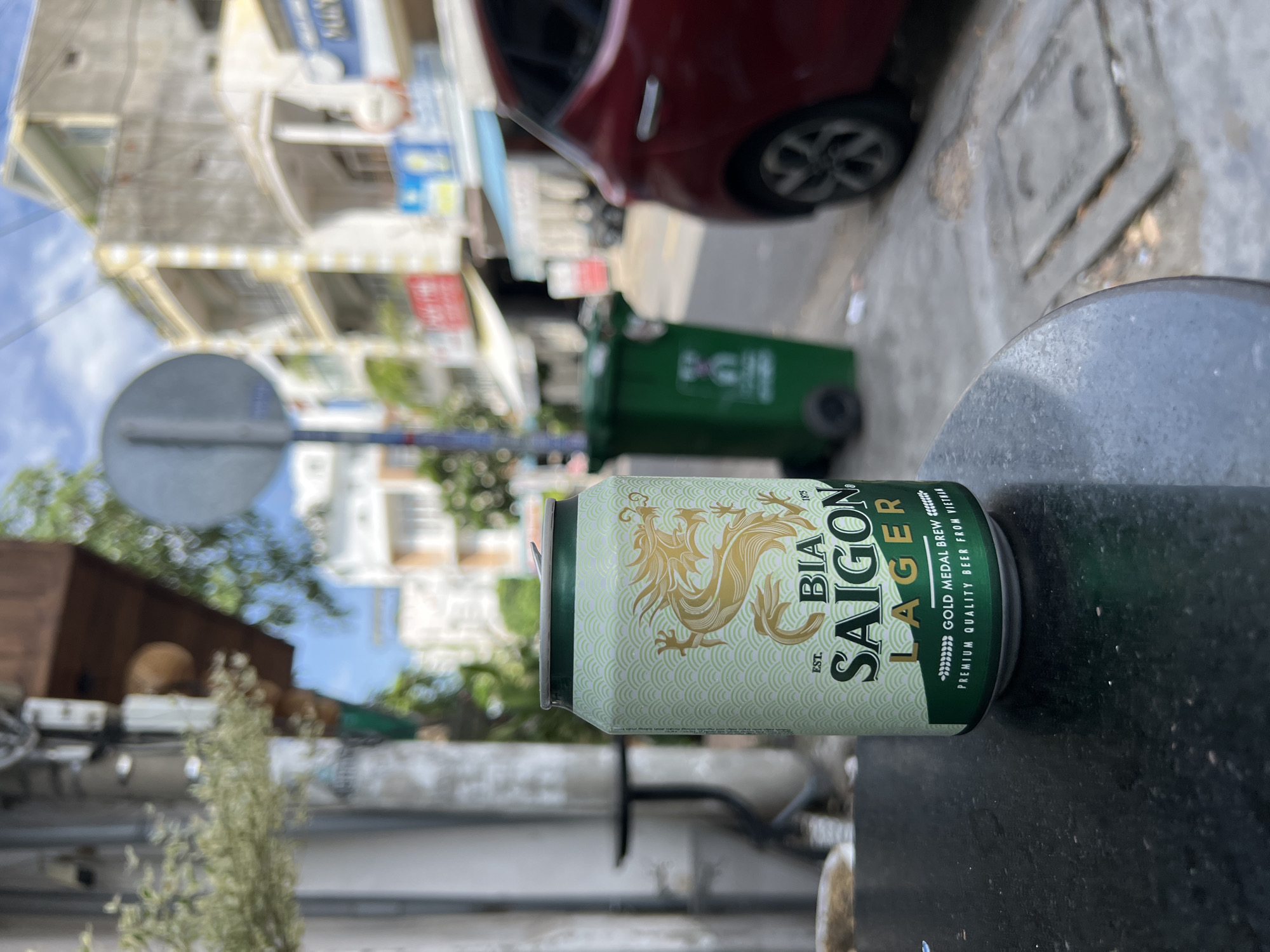 An outdoor shot captures a can of Saigon Lager beer, prominently placed on a dark, speckled table in the foreground. The can features a light green and white design with a golden dragon motif and text indicating "BIA SAIGON LAGER." Behind the can, a blurred street scene unfolds. A red car is parked on the right side of the frame, and a green trash bin stands further back. Buildings with balconies and awnings line the street, typical of urban architecture in Vietnam. The sky above is partly cloudy. The overall setting suggests a street-side cafe or restaurant in Nha Trang, Vietnam, likely during daylight hours. There are no people or discernible actions in the image, presenting a still-life composition of the beer can within its urban environment.