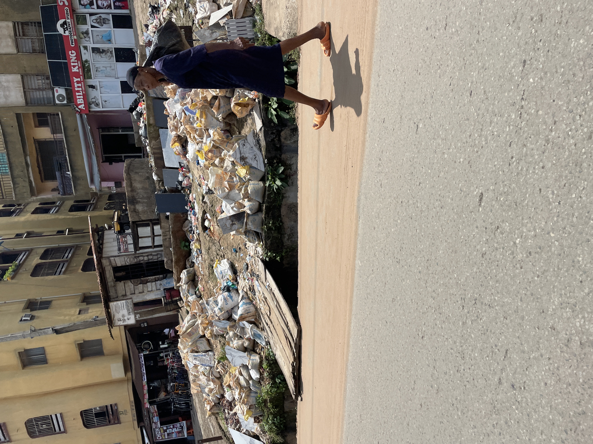 A medium shot captures a woman walking from right to left across a dusty dirt path adjacent to a busy street. She is dressed in a dark blue dress and orange sandals and carries a large black trash bag on her right shoulder. The woman is walking past a large pile of trash bags that have been accumulated along the side of the road. In the background, several buildings can be seen, including a yellow multi-story building and a pink building with a photography studio sign. The street itself is paved, with a strip of dirt and debris separating it from the trash pile and buildings. The sky is bright and sunny, casting shadows on the ground.