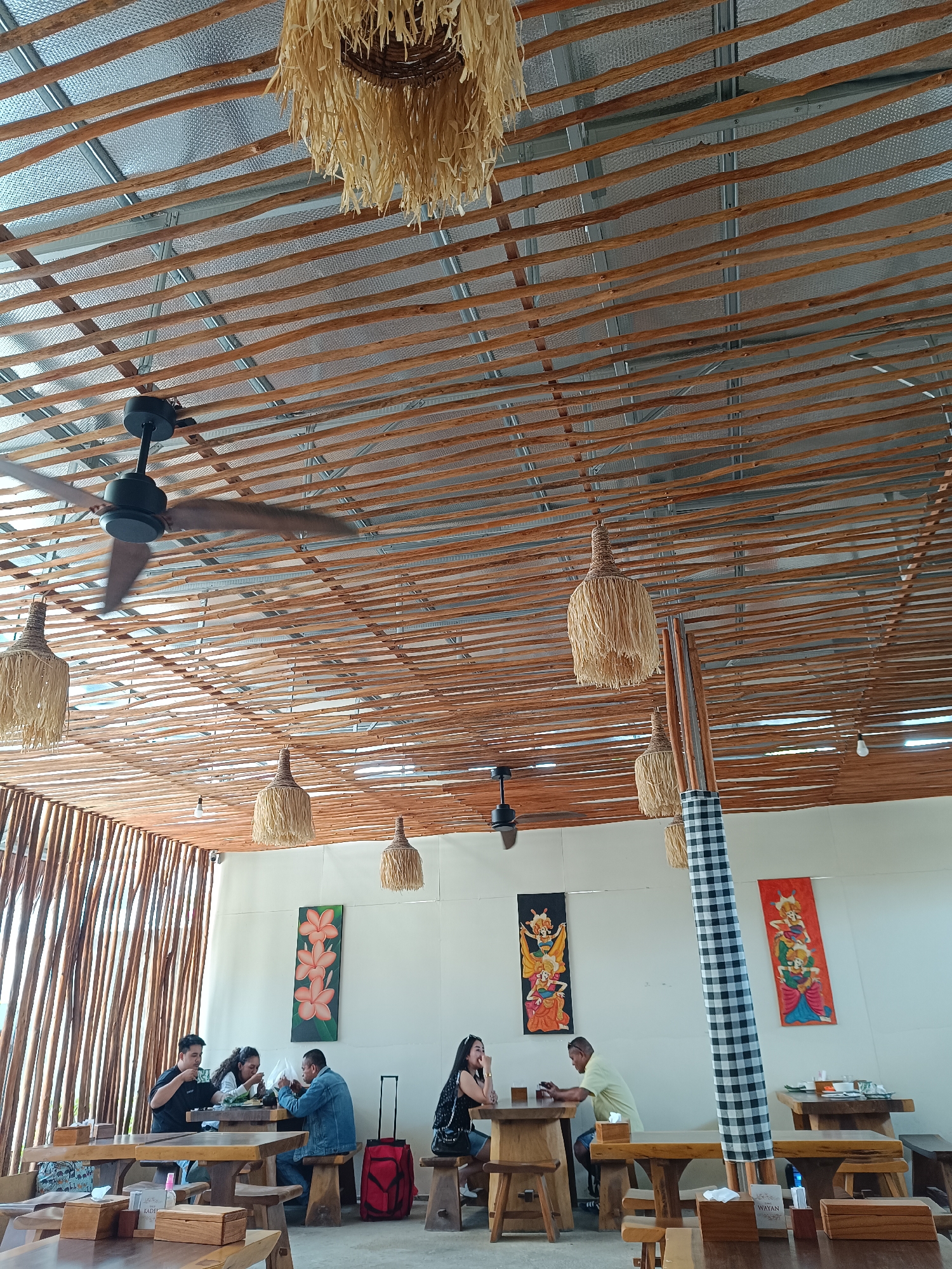 The image depicts the interior of a restaurant with a rustic, natural aesthetic. The ceiling is constructed from numerous parallel wooden beams, creating a slatted effect, with a metallic, textured material visible above the beams. Several pendant lights with woven, straw-like shades hang from the ceiling, along with a black ceiling fan in motion. The walls are white, adorned with colorful, traditional-style paintings featuring figures and floral motifs.

Seated at wooden tables are several individuals. On the left, three people appear to be dining, with one person holding a phone. To the right, two individuals are seated at separate tables, one woman looking away from the camera and another person engaged with their phone. A red suitcase is visible behind the group on the left. The restaurant furniture is predominantly made of wood, with simple wooden chairs and tables. The overall atmosphere suggests a casual dining environment. The presence of decorative elements like the paintings and the ceiling fan suggests it is a place for people to gather.

The scene is located within a restaurant in Sorong, Indonesia.