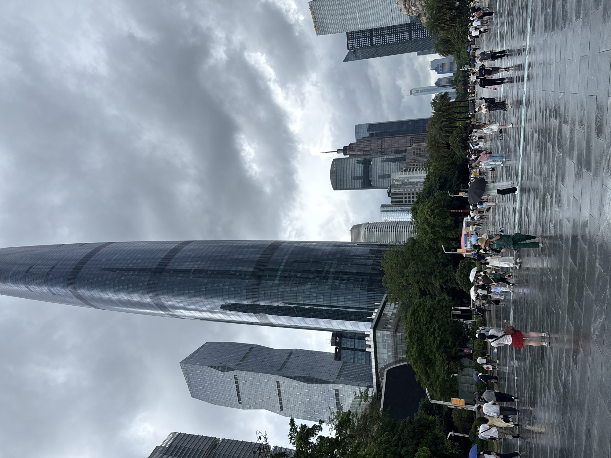 The image depicts a public square in Tianhe District, China, with a cloudy sky overhead. Several modern skyscrapers dominate the background, including a tall, curved glass tower and a twisted geometric building. In the foreground, a wet, paved plaza reflects the overcast sky and surrounding architecture. Numerous pedestrians are present, many holding umbrellas, indicating it is raining. People are walking in various directions across the plaza, some in small groups and others individually. A woman in a red skirt and white top stands facing away from the camera, while a person in a green outfit and straw hat is walking towards the right side of the frame. The scene suggests a bustling urban environment experiencing a rainy day.