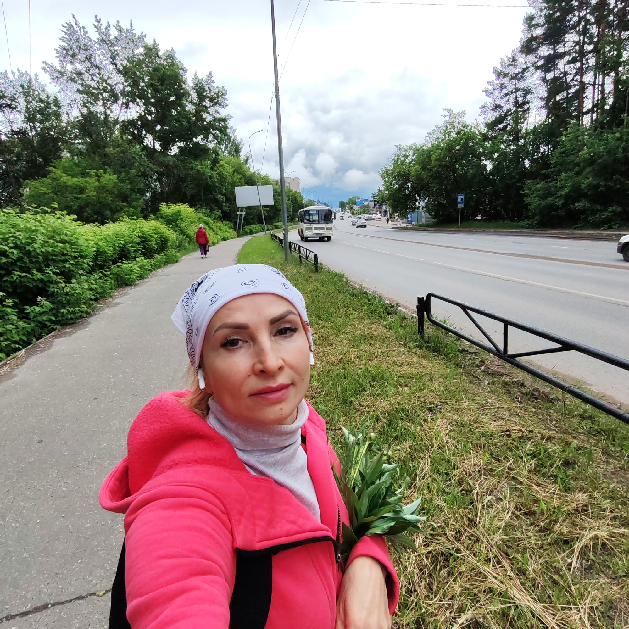 The image depicts an outdoor street scene, identified as being in Chaikovsky, Russia. The setting is an urban or suburban street on an overcast day, characterized by dense vegetation and a mix of human activity and infrastructure.
In the foreground, a woman is taking a selfie. She has fair skin, defined eyebrows, and is looking directly into the camera with a neutral expression. She wears a white bandana with dark patterns, white wireless earbuds in both ears, a light grey turtleneck sweater, and a bright pink long-sleeved jacket with black accents. She holds a bundle of green leaves or plant stems in her left arm, resting against her hip.
To the left of the woman, a grey asphalt or concrete sidewalk extends into the background. A distinct crack or line is visible on the sidewalk surface. The sidewalk is bordered by a continuous line of dense, green deciduous bushes and trees. Further down the sidewalk, another individual, dressed in a red jacket and dark pants, walks away from the camera, carrying a dark bag.
To the right of the woman, a strip of ground features a mix of green grass and dry, straw-like grass. A low, black metal barrier or fence, composed of horizontal bars and triangular supports, runs parallel to the road, separating the grassy verge from the main thoroughfare.
The midground is dominated by a multi-lane asphalt road. A prominent reddish-brown streak is visible horizontally across one of the road lanes. A white or light-colored bus, possibly a PAZ model commonly seen in Russia, travels towards the camera in the rightmost lane. Several other cars are visible further down the road, moving in both directions. A tall, dark grey utility or street light pole stands prominently on the left side of the road. On the far side of the road, a blue rectangular sign with a white pictogram of a person and a bus indicates a bus stop. A large, blank white rectangular sign or billboard structure is also visible on the left side, behind the bus.
The background features numerous tall, green trees, including some with lighter bark, forming a dense canopy on both sides of the road. Distant buildings are partially visible on the horizon through the trees and atmospheric haze. The sky is largely overcast with significant cloud cover, suggesting a cloudy day without direct sunlight.