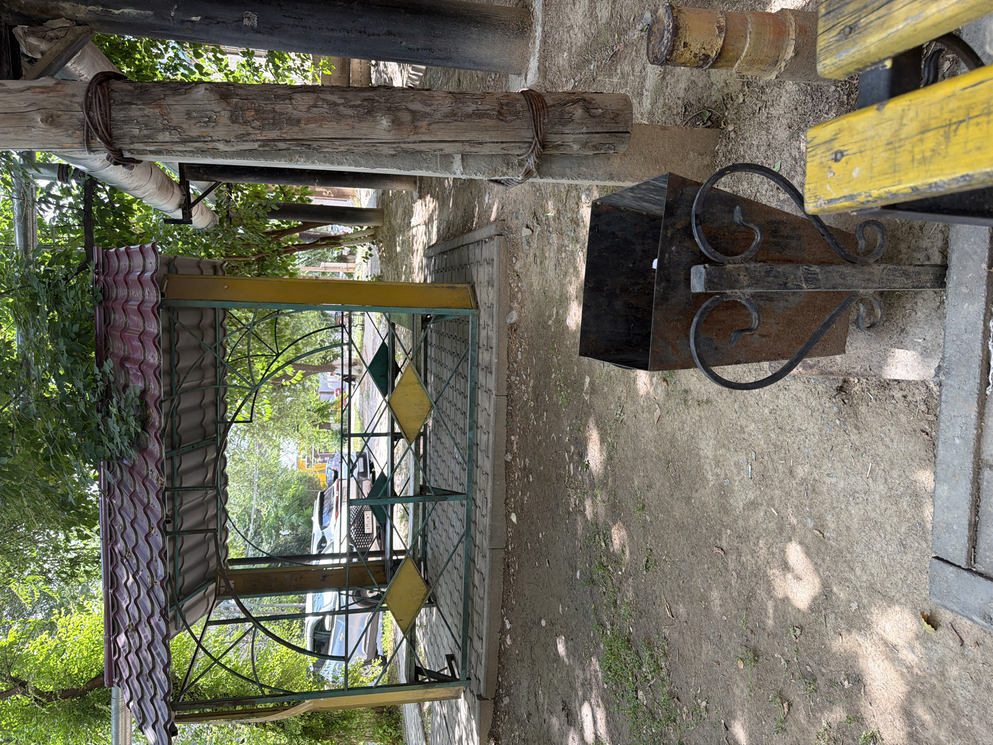 The image depicts an outdoor scene in Shymkent, Kazakhstan, likely a public area or park. In the foreground, a rusty metal trash can with decorative scrollwork rests on a dirt and gravel ground, adjacent to a weathered yellow wooden bench. Behind the trash can, a large wooden post, possibly supporting overhead pipes, is visible. Further back, a covered gazebo with a red tiled roof and green metal railings offers seating. Beyond the gazebo, two white cars are parked on a paved area. The environment is characterized by dirt, scattered leaves, grass, and trees providing shade. The overall impression is one of a functional, somewhat worn, but active outdoor space.