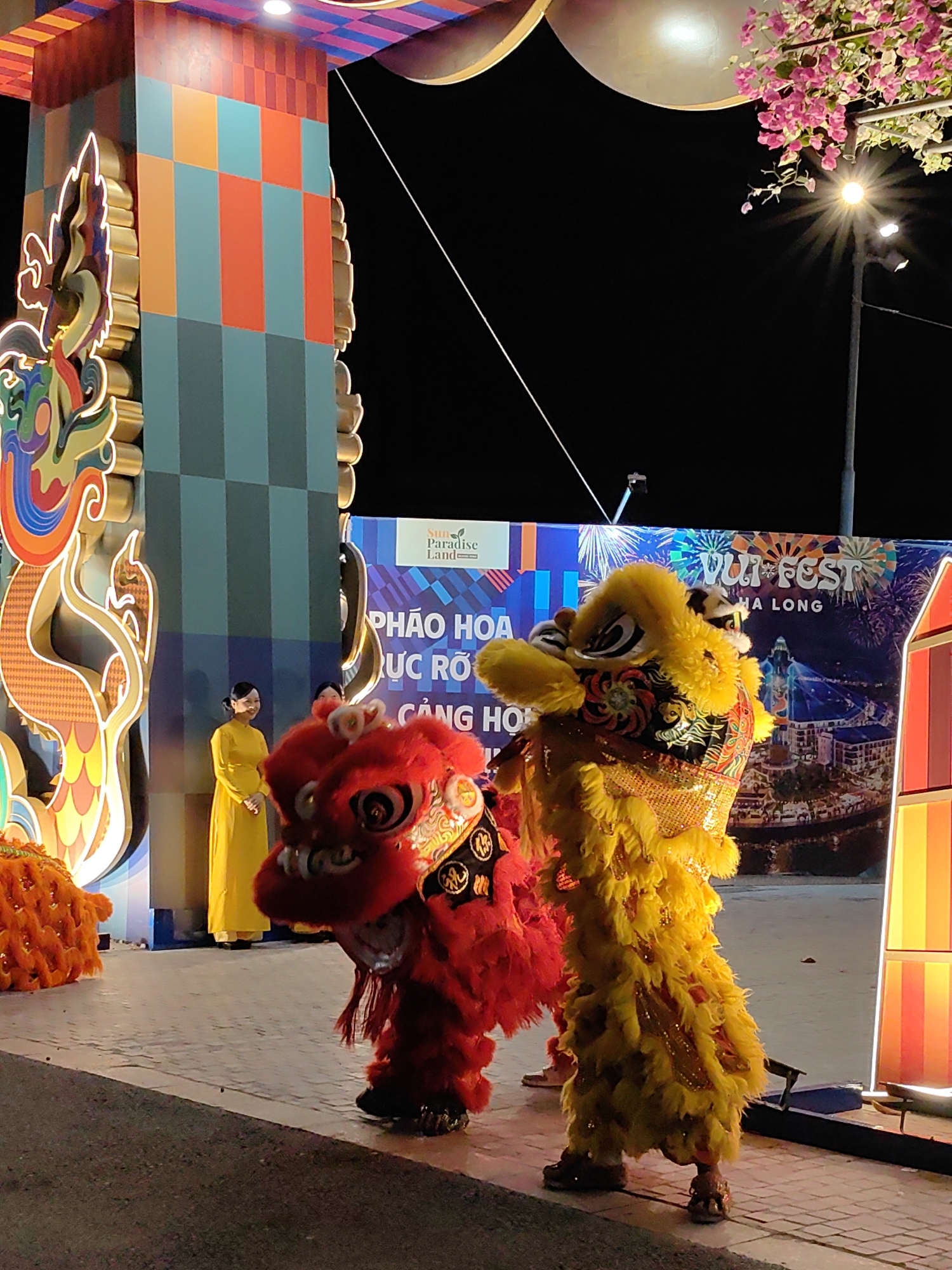 A vibrant nighttime scene in Bai Chay Ward, Vietnam, features two elaborately costumed lion dancers, one red and one yellow, performing on a paved surface. A woman in a yellow traditional dress stands in the background, near a colorful, decorative archway. Behind the dancers is a banner promoting the "Vui Fest Ha Long" event. A street light with pink blossoms hangs nearby. The overall ambiance is festive and celebratory.