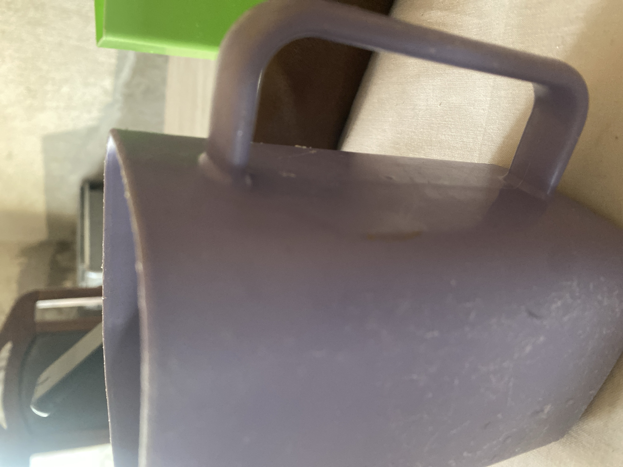 This media file shows a close-up, rotated view of a purple plastic scoop with a handle. The scoop appears to be empty and is resting on a textured, light-colored surface, possibly fabric or a woven material. A portion of a bright green object is visible in the top left corner, which could be another plastic container or item. In the background, out of focus, is a darker, possibly brown or black object, and a hint of a wall or vertical surface. The lighting suggests an indoor setting, possibly during the daytime, with soft shadows. There is no visible text in the image. The focus is on the texture and form of the plastic scoop. The overall impression is one of everyday objects, possibly in a domestic or storage setting.