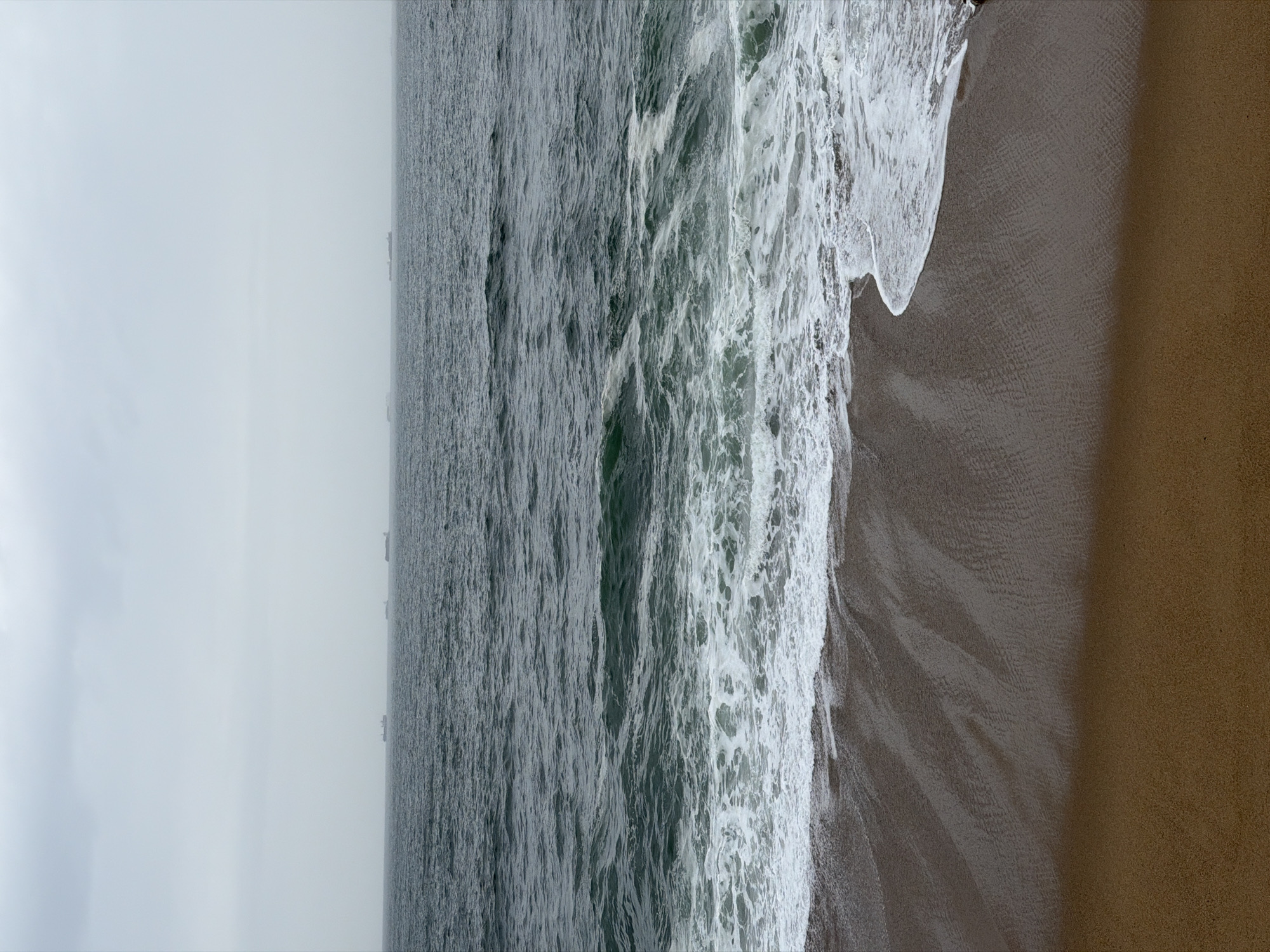 This image shows an aerial view of a beach on a foggy day. The water is rough with white foamy waves crashing onto the shore. The sand is dark and wet near the water, becoming drier and lighter in color further inland. The sky is overcast and pale gray, suggesting a misty or foggy atmosphere. In the distance, there are a series of structures that could be part of a harbor or some other coastal infrastructure. The location is likely a beach in Porto, Portugal, given the coastal geography and architecture sometimes seen on the horizon.