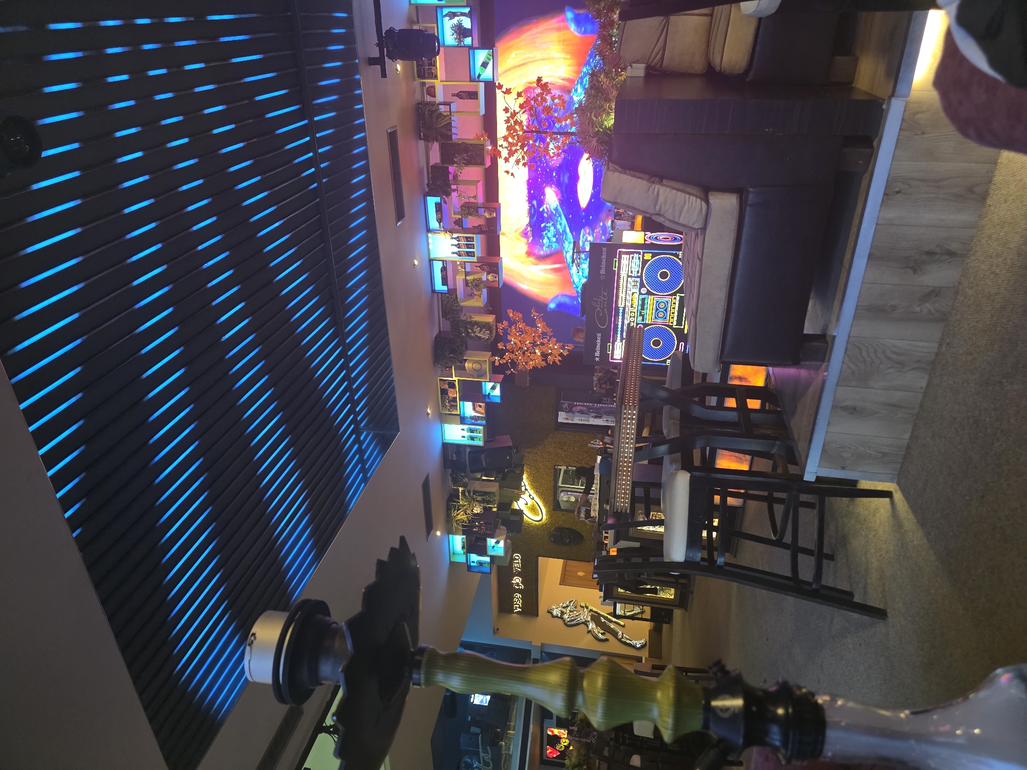 The image depicts the interior of a venue with a modern, eclectic decor. The location is presumably a bar or a lounge, possibly in Sofia, Bulgaria. The ceiling features a series of horizontal panels illuminated with blue LED lights. Various decorative elements are noticeable, including illuminated wall art, plants, and a boombox-styled display. Seating arrangements are present, with sofas and bar stools around tables. The bar area is visible in the background, featuring decorative elements and lighting. A hookah is prominently featured in the foreground.