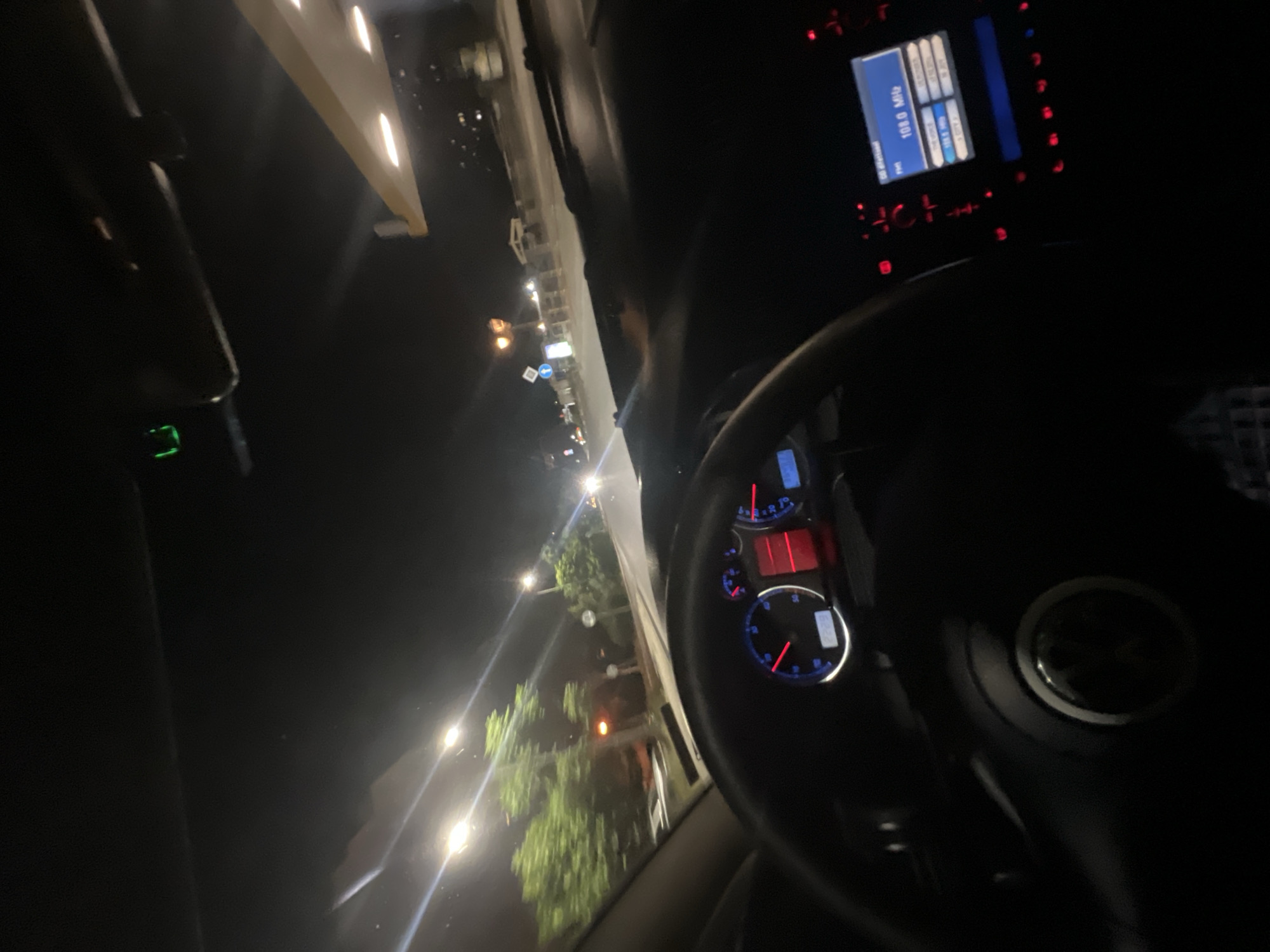 The image captures the interior of a car at night, likely in Plovdiv, Bulgaria, as indicated by the mention of the city. The perspective is from the driver's seat, looking forward. The dashboard is illuminated, showing a speedometer with a needle pointing towards 0, and a red digital display reading "1228." To the right, a car radio display shows "108.0 MHz" and other radio station information. The steering wheel is visible in the foreground, with a car emblem in the center. Outside the car, streetlights illuminate a road and trees. The scene appears to be a typical night drive on a city street, possibly near a gas station, suggested by the brightly lit structure in the upper right portion of the image. There are no people or distinct interactions visible in the frame.