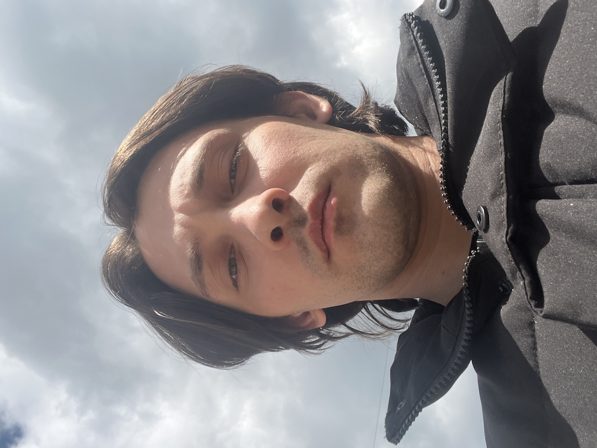 A close-up, low-angle selfie shows a young man with medium-length brown hair, looking directly at the camera. He is wearing a black puffer jacket with a high collar. The background is an overcast sky with visible clouds. The image appears to have been taken outdoors, possibly in the city of Orekhovka, Russia, given the context provided.