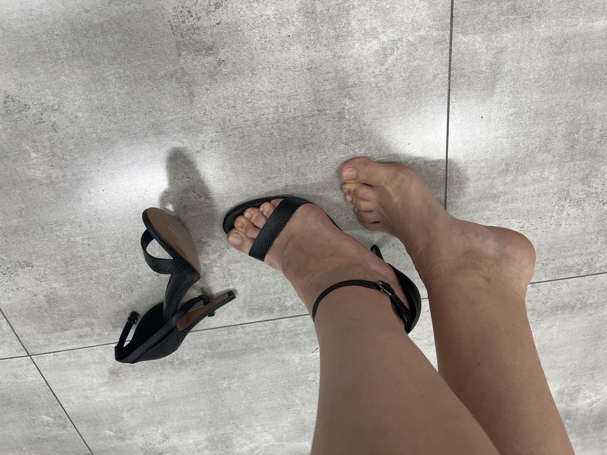 In Afipskiy, Russia, the image captures a close-up, eye-level shot of a person's bare feet and a pair of black high-heeled sandals on a tiled floor. The person's left foot is partially visible, with the right foot adorned by one of the sandals, which features a thin ankle strap and a toe strap. The other sandal lies discarded on the floor to the right. The floor is made of large, light grey tiles with subtle variations in texture and color, possibly resembling concrete or stone. The lighting suggests an indoor environment, casting soft shadows on the floor. The overall scene is static and focuses on the footwear and the feet.