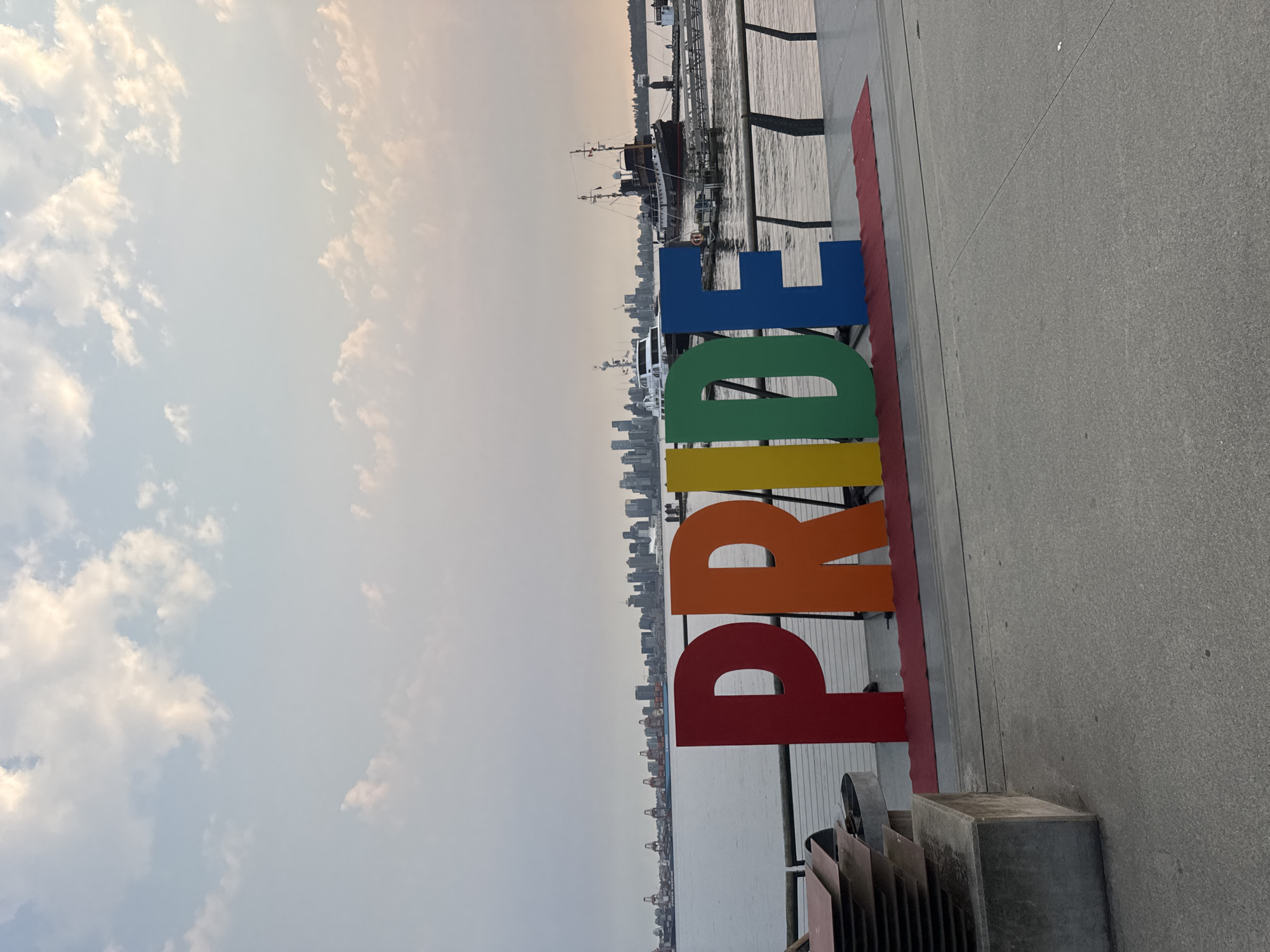 A waterfront promenade in North Vancouver, Canada, features large, colorful, rainbow-colored letters spelling out "PRIDE." The letters are positioned in the foreground on a red carpet. In the background, a calm body of water separates the promenade from a city skyline, which includes numerous buildings and cranes. Several boats are visible on the water, including a tugboat and a ferry. The sky is partly cloudy with a soft, diffused light, suggesting it is either early morning or late afternoon. A concrete barrier with stacked metal sheets is visible to the left of the "PRIDE" installation.