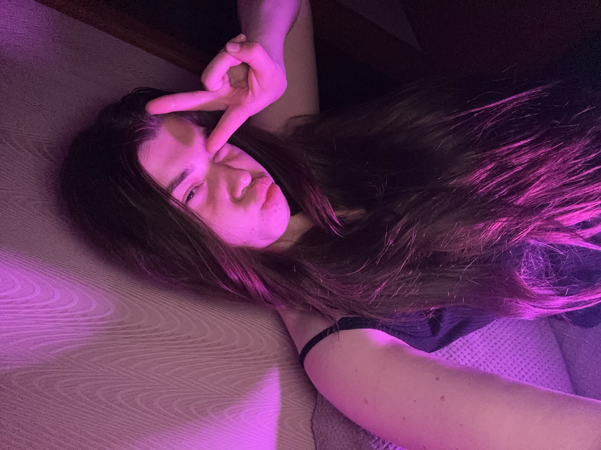 A young woman with long, dark brown hair is captured in a selfie, illuminated by purple lighting. She is leaning against a textured wall, her face turned towards the camera. She is making a "peace" sign with her right hand, her fingers extended near her eye, and her lips are pursed. She is wearing a dark-colored tank top. The background consists of a textured wall and what appears to be a couch or upholstered furniture. The lighting casts a strong purple hue across the scene, particularly on her left arm and hair. There is no discernible urban environment or specific location within Мещёры, Russia, visible in the image.