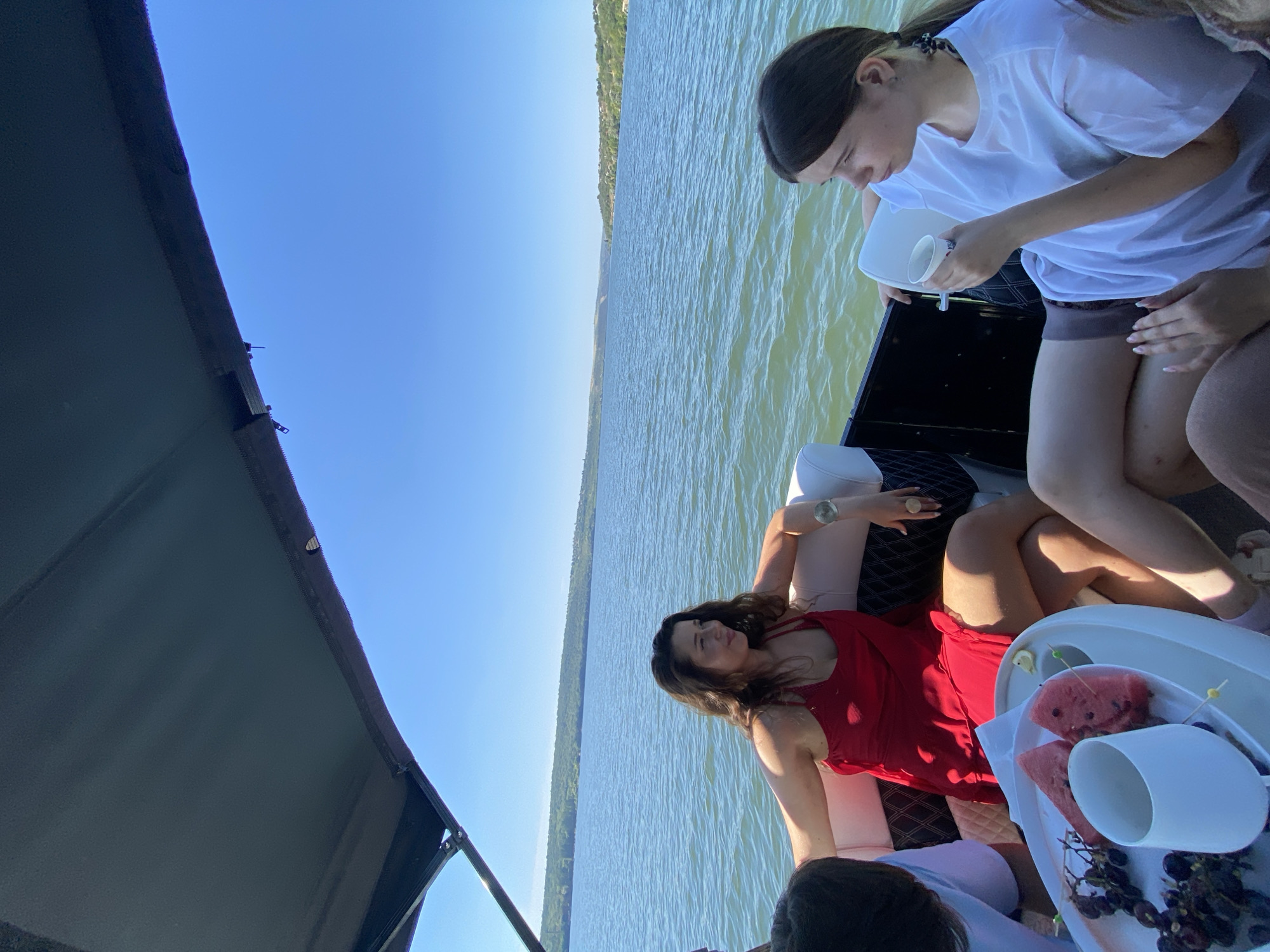 An overhead shot captures a bright, sunny day on the Dniester River near the city of Roghi, Moldova. The scene is set aboard a boat, with a dark canopy providing shade. In the foreground, a woman in a red dress is seated, her left arm resting on the back of the boat. To her right, a young woman in a white t-shirt is holding a white cup. A white tray with watermelon slices, grapes, and a cup is positioned in front of the woman in the red dress. The river stretches out into the distance, with a tree-lined shore visible on the horizon. The sky is a clear, vivid blue.
