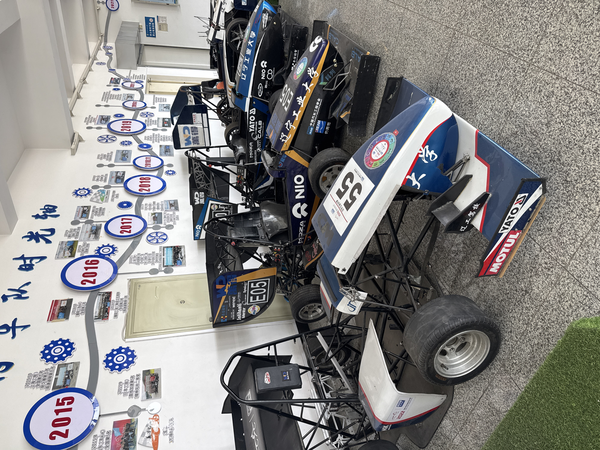 The image shows a room with a timeline on the wall displaying years 2015 to 2021, labeled "牛队时光轴" (likely translated as "Ox Team Timeline"), and several open-wheel race cars lined up along the wall. The timeline includes photos and text written in Chinese detailing the achievements and history of a team. The room has a tiled floor and fluorescent lighting. The race cars vary in design but share similar open-wheel chassis. Logos and names like "YATO," "MOTUL," "NIO," "辽宁工业大学" (Liaoning University of Technology), and "WD" (likely representing Western Digital) are visible on the cars, suggesting sponsors and the university affiliation of the team. The car nearest to the viewer has a large number "55" on it. The timeline details various race achievements. The environment indicates this may be an engineering or automotive laboratory or display area affiliated with Liaoning University of Technology in the city of Zhongtun, China, but the specific location in the city cannot be verified.